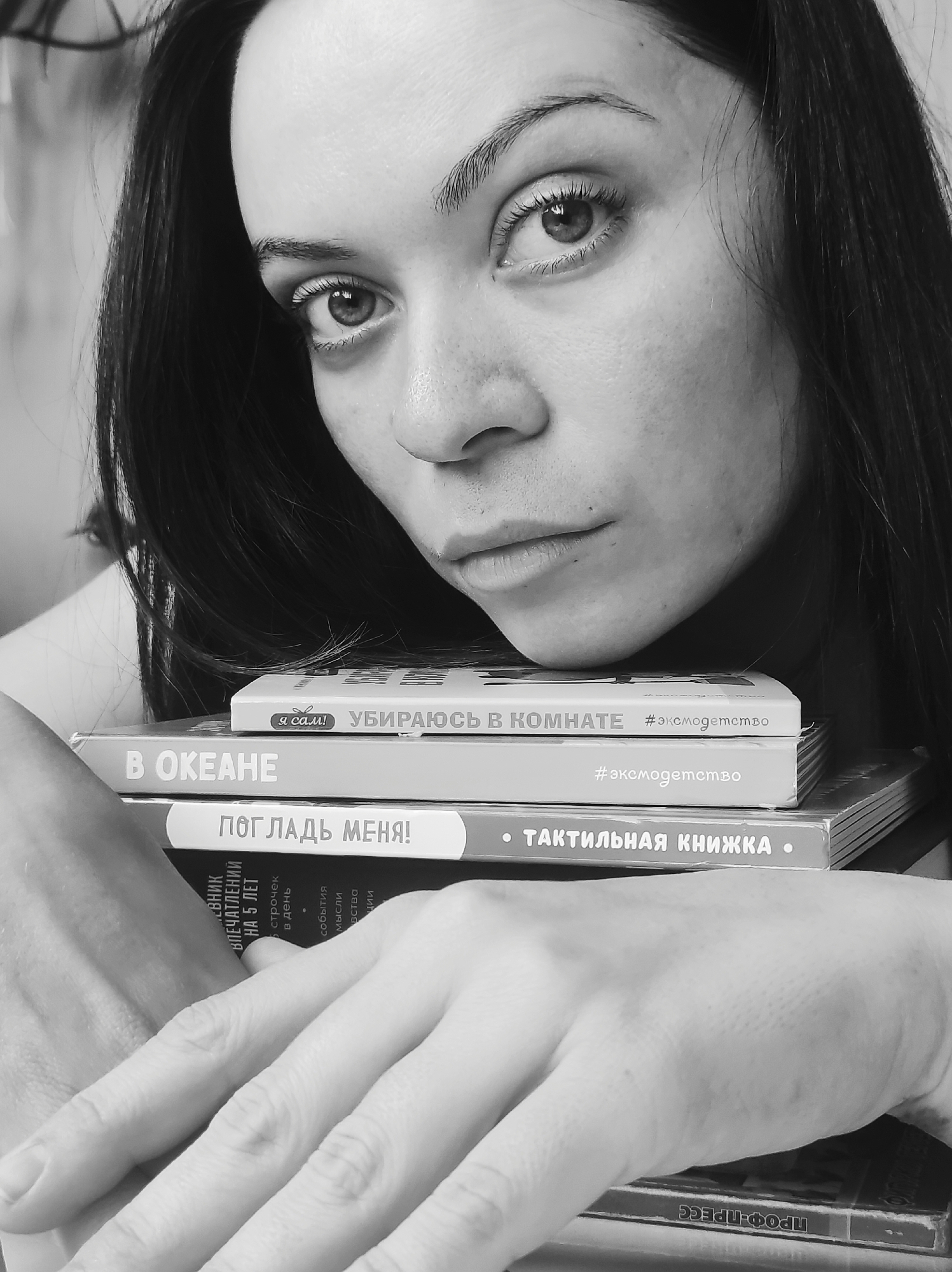 A black and white close-up portrait shows a woman with long dark hair, her head resting on a stack of books. She looks directly at the camera with a serious, contemplative expression. Her dark hair frames her face, and her eyebrows are well-defined. Her hands are also resting on the books, one on top of the other, near the bottom right of the frame.
The central activity is the woman posing with the books, suggesting an interest in reading or a connection to the subjects of the books. The setting appears to be indoors.
Notable details include the texture of her skin and the details of her eyes, which are sharply in focus. The monochrome palette adds to the serious tone of the image.
Visible text on the books, from top to bottom, includes:
* "я сам! УБИРАЮСЬ В КОМНАТЕ" (I myself! TIDY UP THE ROOM) with the hashtag "#эксмодетство" (#exmodetstvo).
* "В ОКЕАНЕ" (IN THE OCEAN) with the hashtag "#эксмодетство" (#exmodetstvo).
* "ПОГЛАДЬ МЕНЯ! • ТАКТИЛЬНАЯ КНИЖКА •" (PET ME! • TACTILE BOOK •).
* A book with partially visible text, likely a diary: "ЕВНИК ВПЕЧАТЛЕНИЙ НА 5 ЛЕТ" (DIARY OF IMPRESSIONS FOR 5 YEARS), "5 строчек в день" (5 lines a day), "события Мысли" (events Thoughts).
* Another book at the very bottom shows "ПРОФ-ПРЕСС" (PROF-PRESS), likely a publisher's name.