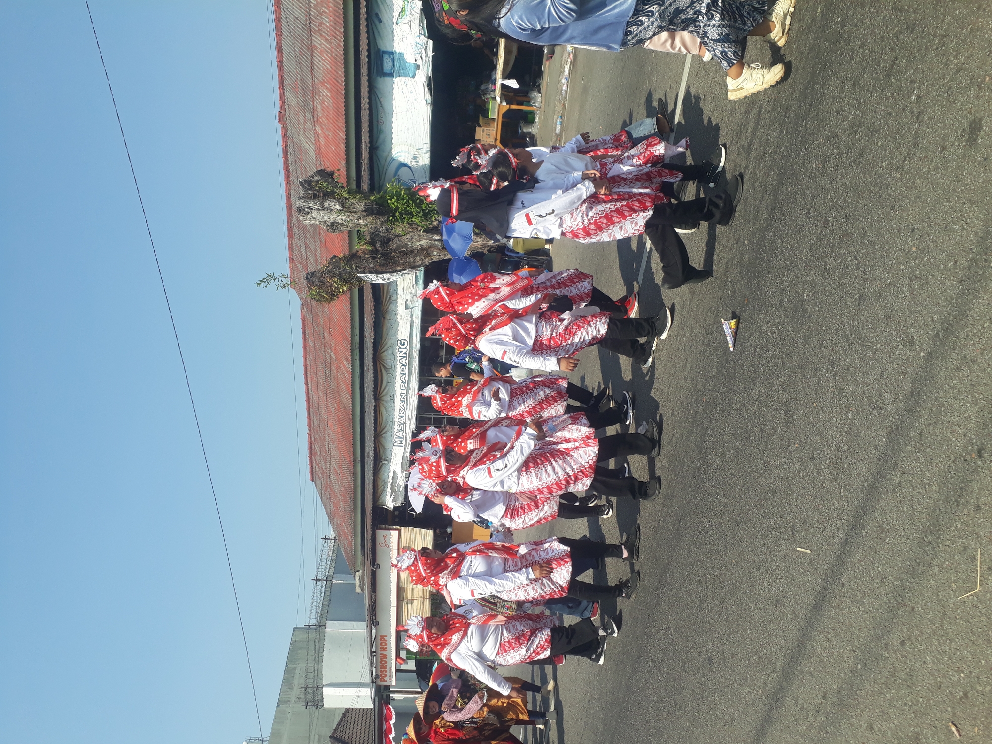 In Pangkalpinang, Indonesia, a group of people in matching red and white traditional attire are participating in a parade or cultural event. The individuals are walking down a paved road under a clear blue sky. The attire includes white tops with red trim, red and white patterned skirts or sarongs, and red headscarves adorned with white feathered headpieces. Some individuals are holding small white umbrellas. The background features buildings with red corrugated roofs, one of which has signage indicating "POSKOW KOPI" and "MASAKAN PADANG". The scene captures a vibrant cultural procession in an urban setting.