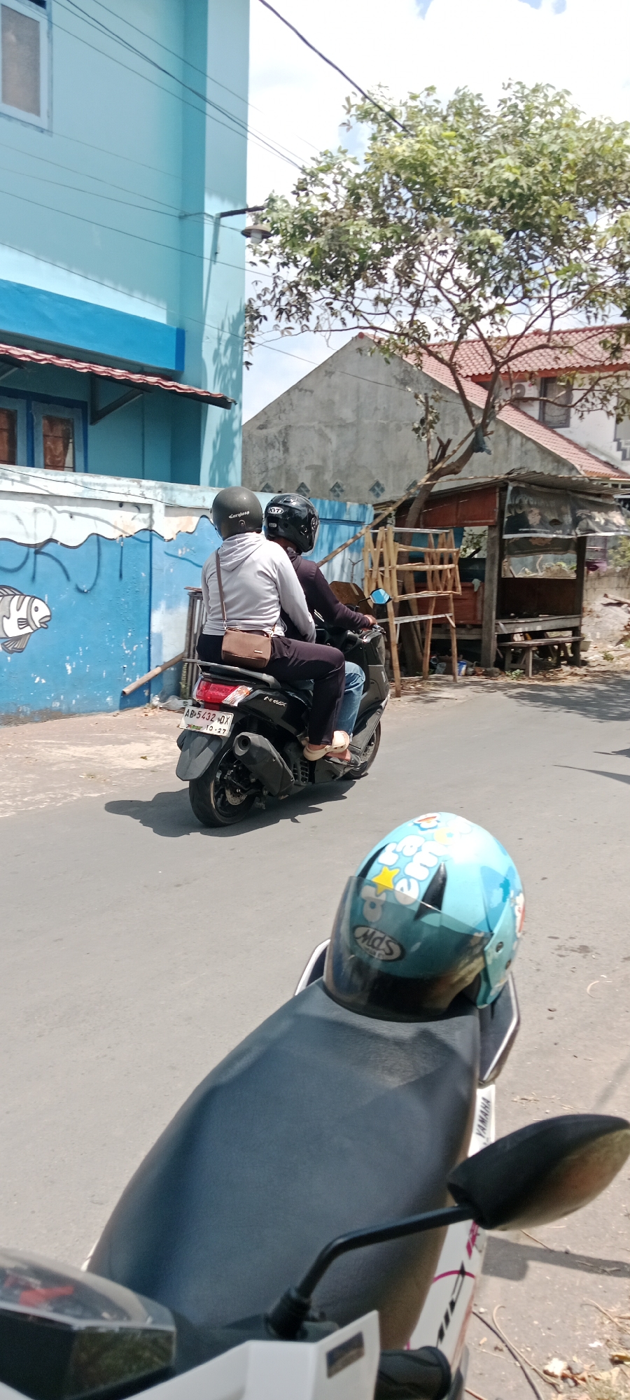 In Purwomartani, Indonesia, two individuals are riding a scooter on a paved road. The person in the front is wearing a dark helmet and a dark long-sleeved shirt, while the passenger in the back is wearing a dark helmet and a light grey hooded sweatshirt, with a brown crossbody bag. The scooter is black, with a license plate that reads "AB 5432 DX 10-27". In the foreground, a light blue helmet with cartoon characters and the brand "Mds" is placed on the back of a white scooter. The background features a light blue building with a white-framed window, a mural of a fish on a blue wall, and a tree with green foliage. Further in the background, there is a simple structure with a red-tiled roof and wooden scaffolding. The sky is bright and cloudy.