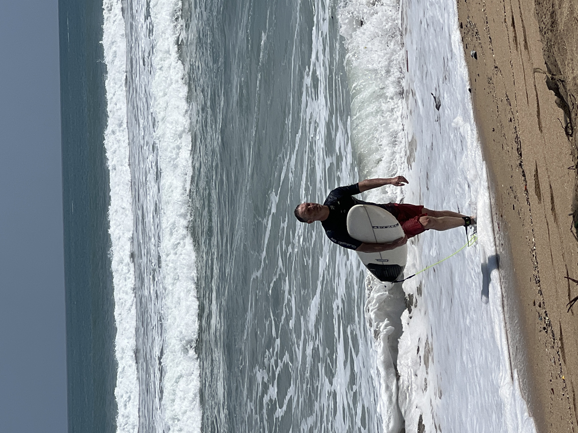 A middle-aged Caucasian male wearing a black wetsuit top and red board shorts is walking out of the ocean, carrying a white surfboard under his right arm. The surfboard has a leash attached to his ankle. He is walking on a sandy beach with waves breaking around his legs. The ocean is a deep blue-green with white surf. The sky is overcast. The location is the Kuta beach in Bali, Indonesia.