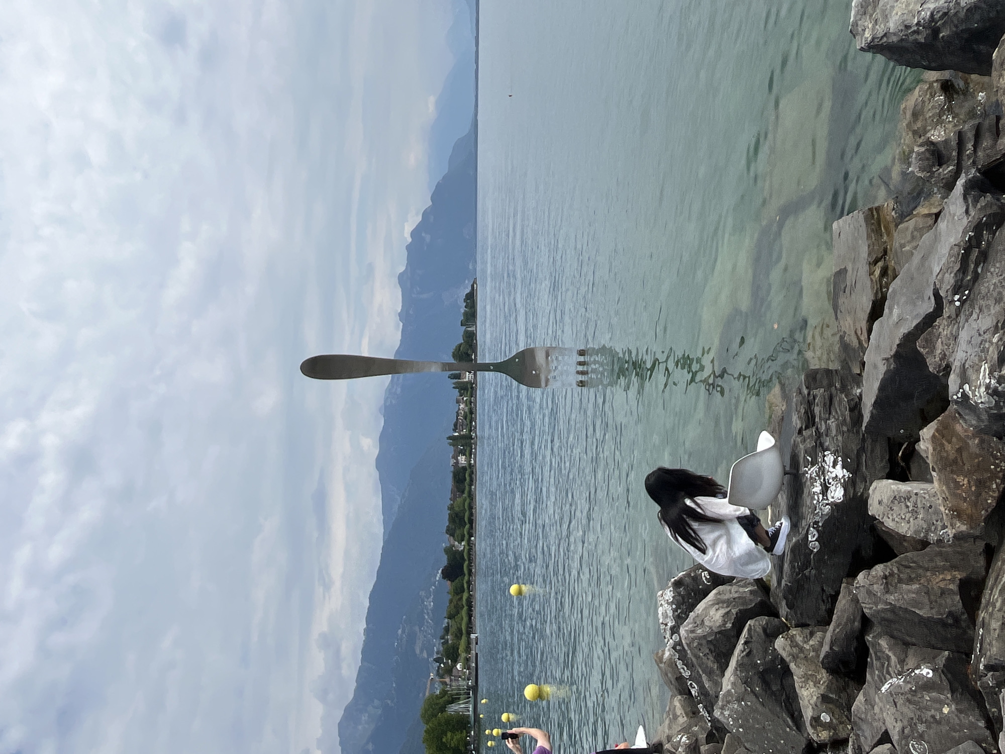 The image depicts a large, stainless steel fork sculpture standing upright in the water of Lake Geneva, in Vevey, Switzerland. The sculpture is positioned in the foreground, partially submerged, with its tines extending upwards. In the middle ground, a young woman with long dark hair, wearing a white shirt and dark pants, is crouched on a rocky shore, reaching towards a small white plastic stool. To the left, another person in a purple shirt is visible, holding a phone and taking a picture. Several yellow buoys are scattered across the surface of the lake. The background features a shoreline with trees and buildings, and beyond that, a range of hazy blue mountains under a cloudy sky. The water is clear and turquoise, revealing the lakebed in some areas.