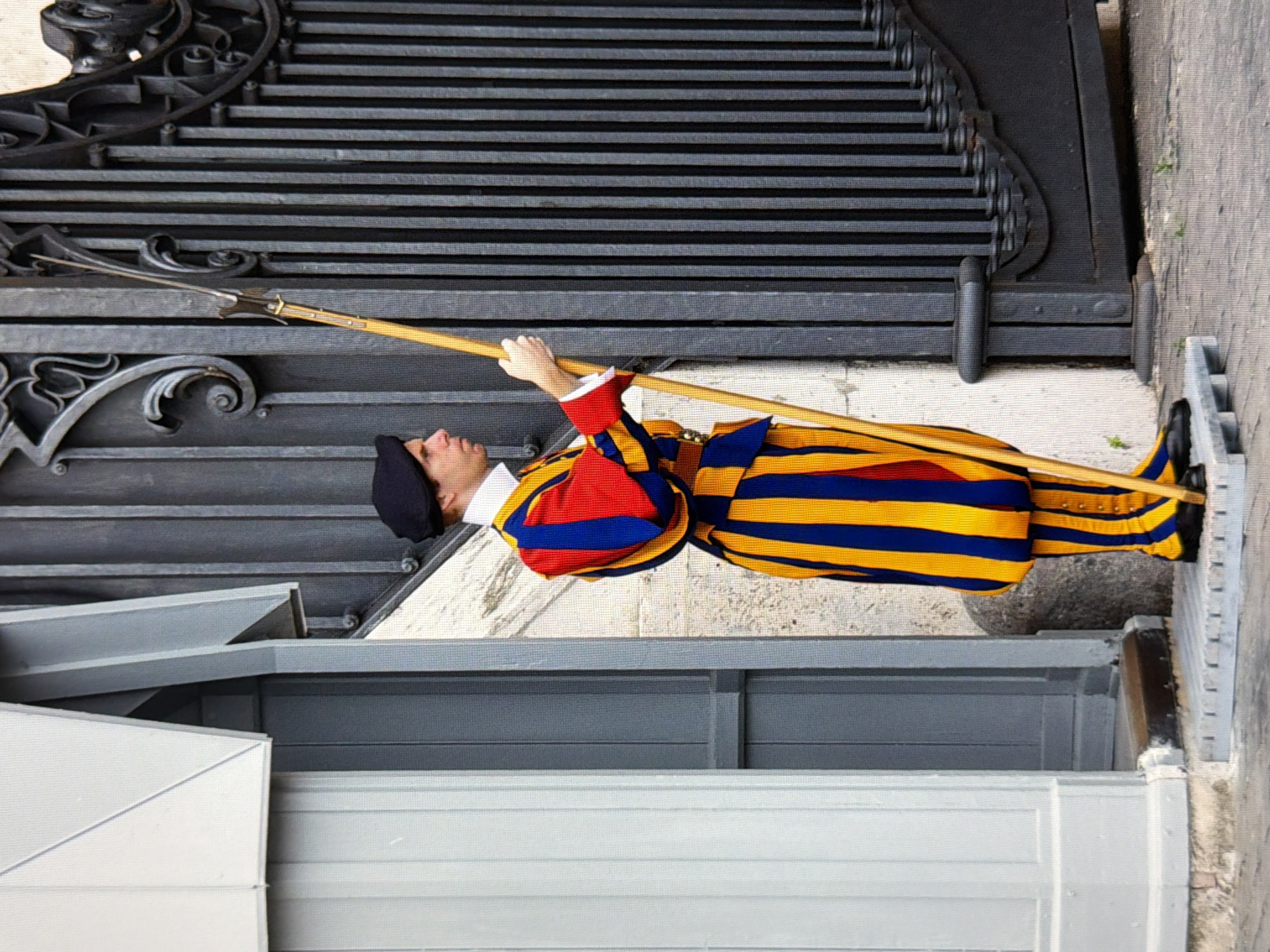 A Swiss Guard stands at attention in front of a dark metal gate. The guard is wearing a uniform of red, blue, and yellow stripes, with a black beret and black shoes. He is holding a halberd in his right hand. The gate is ornate, with decorative scrollwork. To the left of the guard is a light-colored building with white and gray panels. The ground is paved with gray cobblestones. The lighting suggests it is daytime. This image was captured in Kyiv, Ukraine.