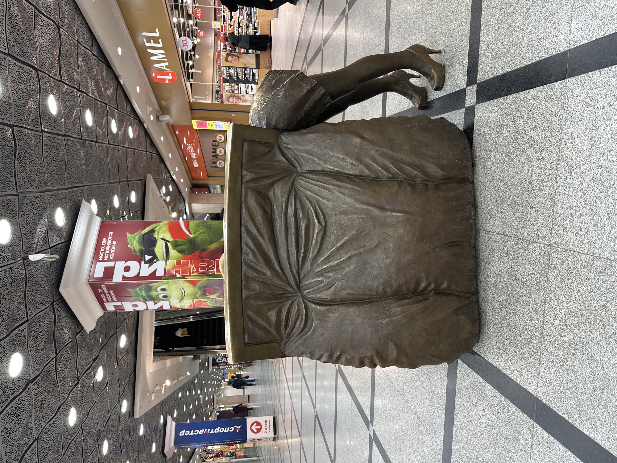 The image captures an indoor scene, likely a shopping mall in Yekaterinburg, Russia, featuring a prominent bronze statue. The statue depicts the lower half of a human body, specifically legs and hips clad in a skirt and high-heeled shoes, appearing to dive head-first into or emerge from a large, rectangular, crumpled bronze form resembling a bed or cushion.
In the background, typical mall elements are visible. The floor is tiled with light speckled patterns intersected by dark stripes. The ceiling features a dark, wavy, perforated design with numerous recessed lights. Various storefronts line the passage, displaying merchandise. Blurry figures of people can be seen walking in the distance, indicating an active commercial environment.
Visible text includes:
- "ЛАМЕЛ" (LAMEL), likely a cosmetics or beauty store, with signs for "Мужская линия" (Men's line) and "Зубные па" (Toothpaste), as well as discount percentages like "-30%" and "-40%".
- A large poster for "ГРИНВИЧ" (GRINVICH), possibly the name of the mall or a major store, with the slogan "место, где исполняются желания" (a place where wishes come true).
- "спортмастер" (Sportmaster), a popular sporting goods chain.
- Directional signage that reads "2 этаж 4 очередь" (2nd floor, 4th section/queue).
- A slogan "красота-уход-забота" (beauty-care-well-being).
The image is captured under artificial lighting, suggesting it could be daytime or evening indoors. The whimsical nature of the statue adds an artistic touch to the commercial setting.