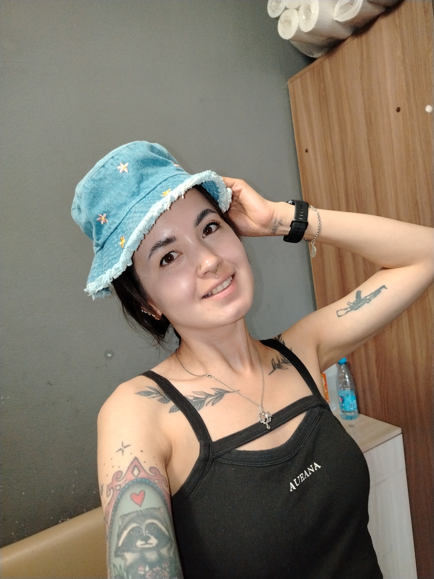 A person with tattoos on their arms and chest is smiling and looking towards the camera, wearing a blue bucket hat decorated with embroidered flowers. They are wearing a black tank top with the word "AUEANA" on it. They are holding the hat with their right hand. The background consists of a gray wall and a wooden cabinet, with rolls of paper on top. A water bottle is visible on the cabinet. There is no mention of the location in the city of Noviy, Russia.