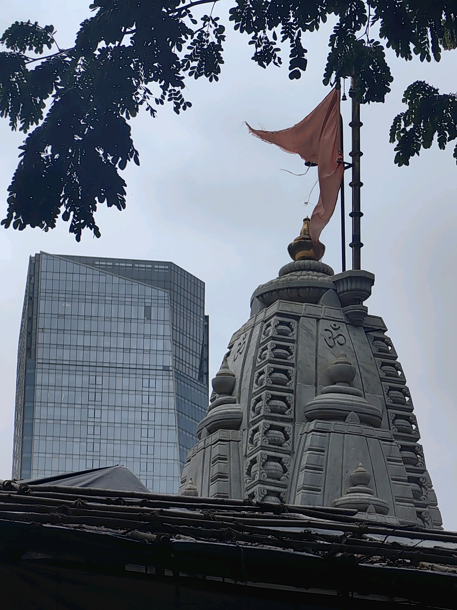 The image captures a view of the Signature Building in Bandra, Mumbai, India. The building is a modern skyscraper with a glass facade and a distinctive angular design. In the foreground, a Hindu temple spire is visible, adorned with intricate carvings and the "Om" symbol. A saffron-colored flag flies from a flagpole atop the temple. Silhouetted branches of a tree frame the top of the image, contrasting with the stark modern architecture of the skyscraper. The sky is overcast, creating a diffused light that softens the scene. A rustic, makeshift roof structure occupies the bottom portion of the frame, providing a textured foreground that emphasizes the juxtaposition of old and new in the urban landscape.