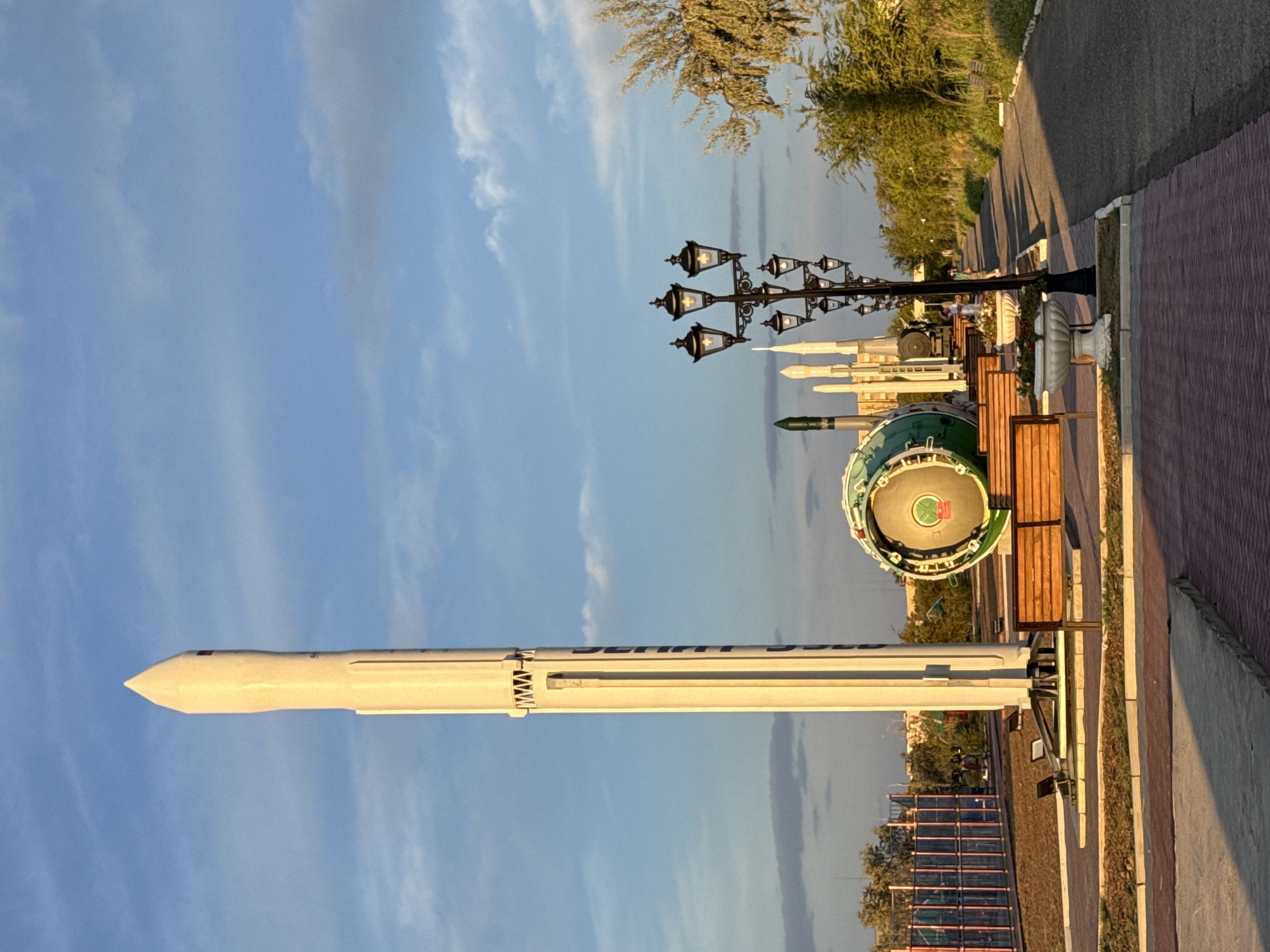 The image depicts an outdoor display of rockets and space-related artifacts within a park-like setting in Baikonur, Kazakhstan. A tall, white rocket stands prominently in the foreground, positioned on a metal stand. To its right, a large, green, cylindrical object, possibly a rocket engine or capsule, is visible. In the background, several other rockets of varying sizes and designs are displayed, some appearing as models or replicas. A lamppost with multiple lights stands to the right of the rockets. Benches are placed in front of the display, suggesting a viewing area for visitors. A paved walkway runs alongside the display, bordered by greenery. In the far background, a playground structure with colorful fencing is visible, indicating the park's use as a recreational space. The sky is partly cloudy, with a mix of blue sky and soft white clouds. The overall scene suggests a day with pleasant weather, possibly late afternoon given the lighting.