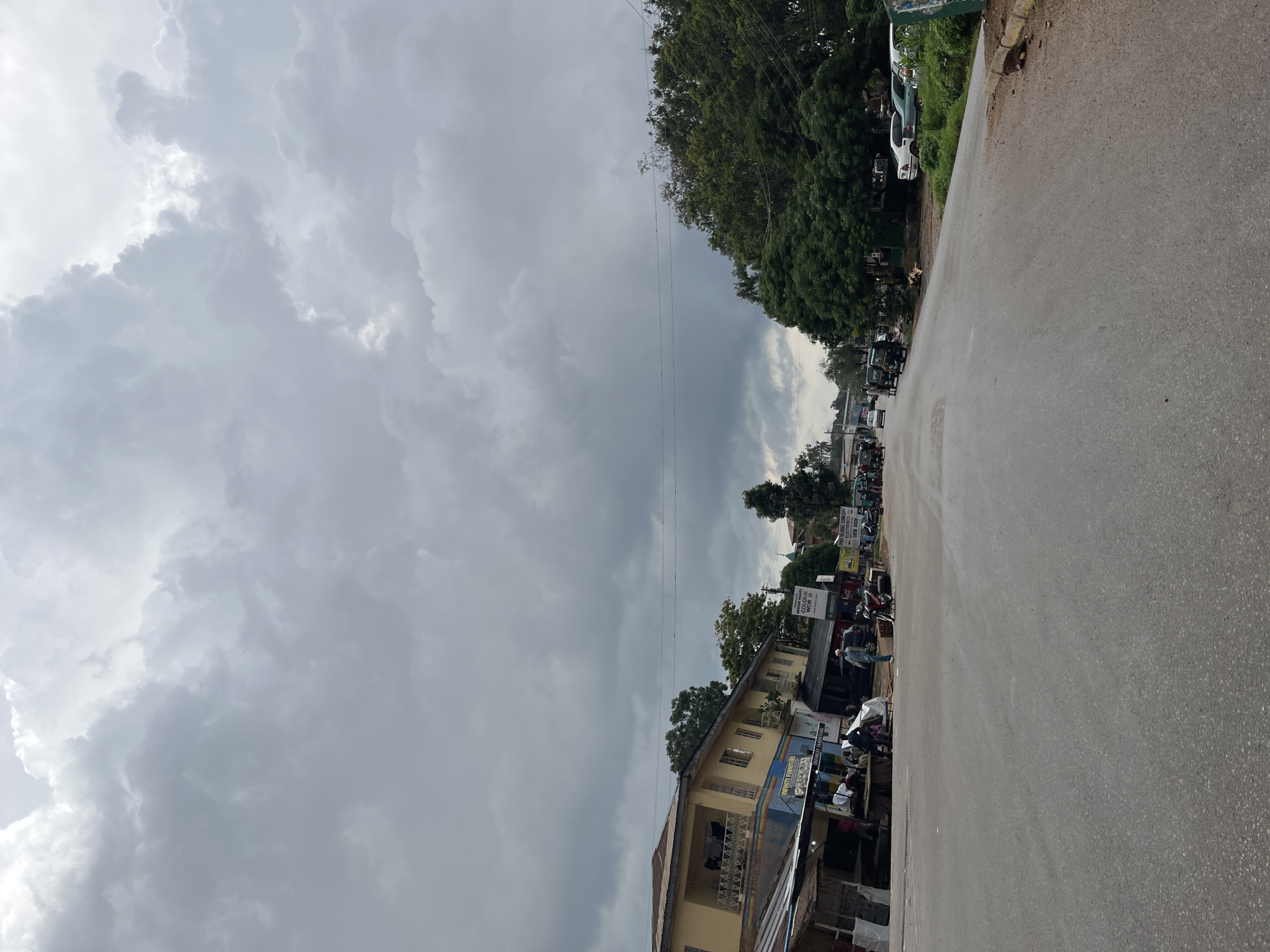The image depicts a street scene in Jos, Nigeria, under a heavily overcast sky with dramatic cloud formations. The road, paved and appearing wet, stretches into the distance. On the left side of the road, a two-story building with a yellow facade and balconies stands, adjacent to a row of shops with corrugated iron roofs. Signs for "INFINITY FURNITURE" are visible. Several people are gathered in front of these shops, some seated. A few motorcycles and a car are parked nearby.
On the right side of the road, lush green trees form a dense barrier. Several vehicles, including cars and what appear to be auto-rickshaws, are parked along the roadside. The overall impression is of a moderately busy street scene, possibly on the outskirts of a town or in a less densely populated urban area, with the impending weather contributing to a subdued atmosphere.