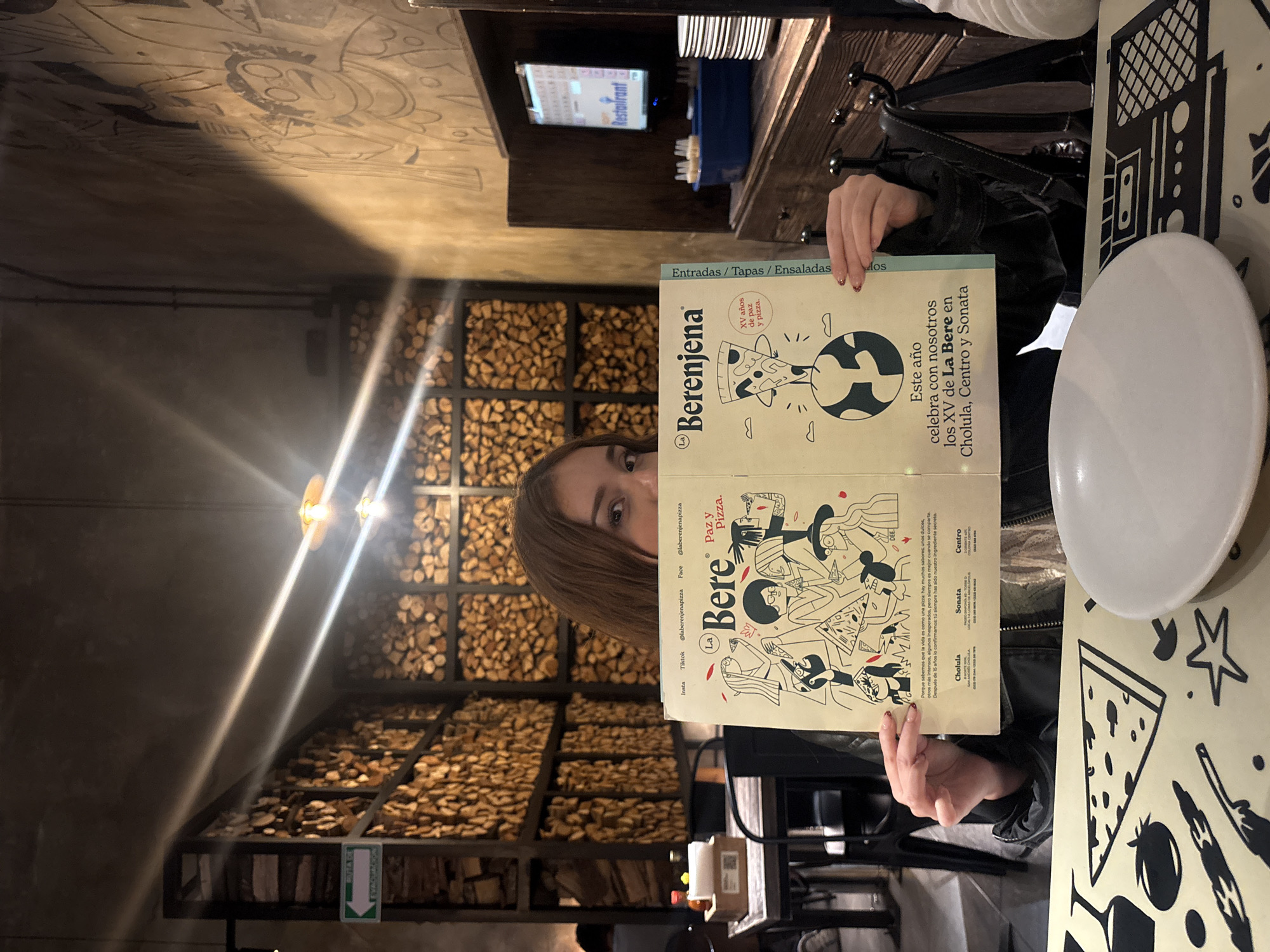 The image displays the interior of a "La Berenjena" pizza restaurant, identified by its menu, located in San Bernardino Tlaxcalancingo, Mexico. A person is seated at a table in the foreground, holding an open menu that largely obscures their face; only their eyes, forehead, and hands with painted nails are visible. The menu is titled "La Berenjena" and "Paz y Pizza," features cartoon illustrations, and references "XV años de paz y pizza" with locations including Cholula. An empty white plate rests on the table, which is covered by a placemat with black graphic designs of food items and geometric shapes.
In the background, a decorative wall composed of dark metal frames filled with stacked firewood is prominent. To the left of this wall, a green emergency exit sign, reading "RUTA DE EVACUACIÓN" with a left-pointing arrow, is affixed to a dark structural column. To the right of the seated individual, a rustic wooden cabinet or counter holds a blue tray containing multiple small, white cylindrical objects, and a stack of white plates. Above this cabinet, a screen displays the word "Restaurant" in blue lettering. The far-right wall features dark, cartoonish mural art. The lighting is warm and artificial, causing visible lens flares in the photograph. A second individual is partially discernible in the distant background, seated at another table, obscured by the firewood wall.