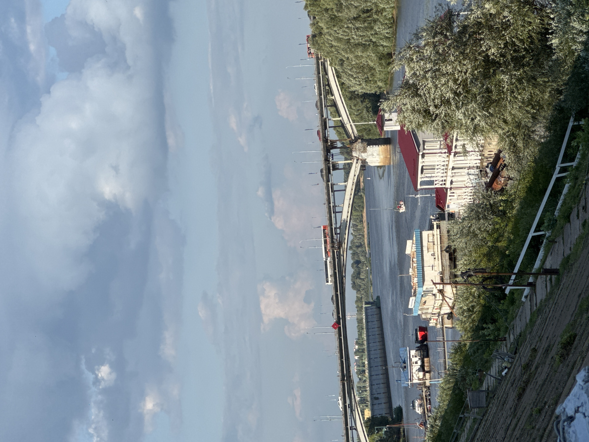 The image shows a wide river with a large bridge spanning it, possibly the Kanavinsky Bridge in Nizhny Novgorod, Russia. Cars and a bus are visible on the bridge. Along the riverbank, there are several boats and barges, some of which appear to be moored. A large building with a red roof, resembling a boathouse or a recreational facility, is situated by the water. The riverbank in the foreground is sloped and covered with vegetation and a path. The sky is partly cloudy with a mix of blue and white. The scene suggests a daytime view of a busy river port or waterfront area in a city.