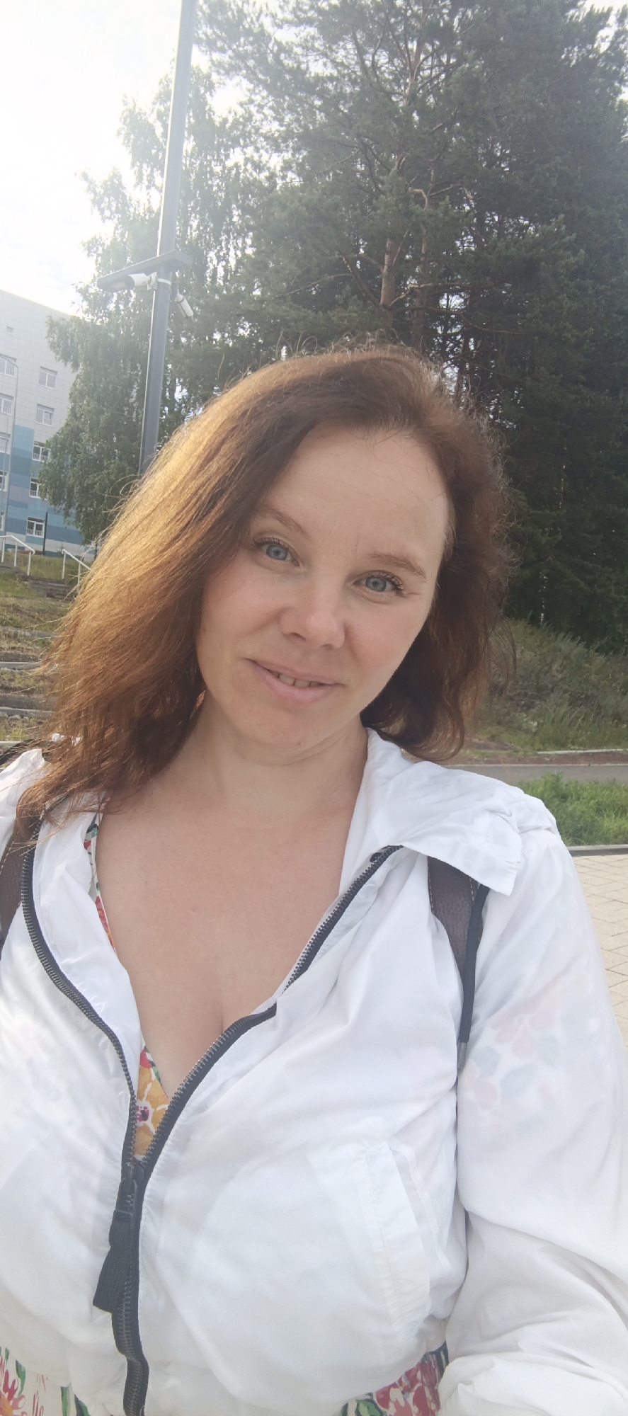 A woman with shoulder-length reddish-brown hair and blue eyes smiles at the camera, looking slightly to her right. She is wearing a white zippered jacket over a colorful floral top. The background shows trees and a building, suggesting an outdoor setting in a somewhat urban or suburban area. The lighting indicates it is daytime, with the sun creating a slight lens flare and highlighting her hair. A lamppost with two security cameras is visible to her left, and a paved walkway is in the foreground. The scene appears to be in Chaikovsky, Russia, and the overall mood is relaxed and pleasant.