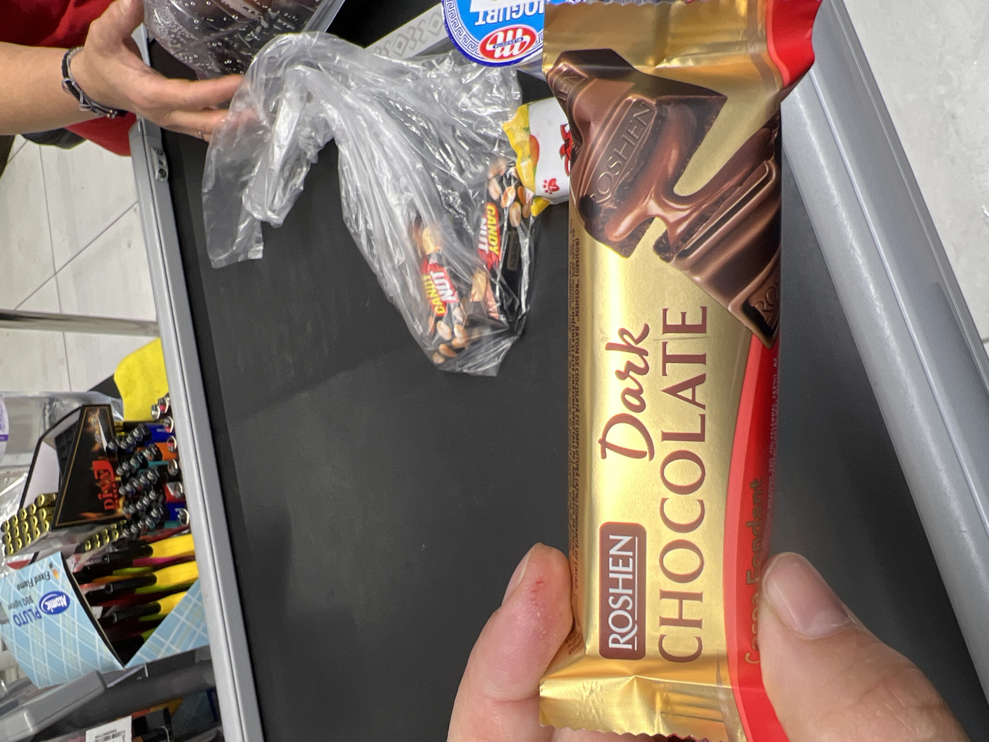 A hand is holding a gold-wrapped ROSHEN Dark Chocolate bar over a black conveyor belt. Next to the chocolate bar, a plastic bag contains two "CANDY NUT" bars. To the right, a blue and white yogurt cup is visible. In the background, a display case holds several lighters and pens. The overall scene appears to be a retail checkout counter, likely in Chișinău, Moldova, given the product branding.