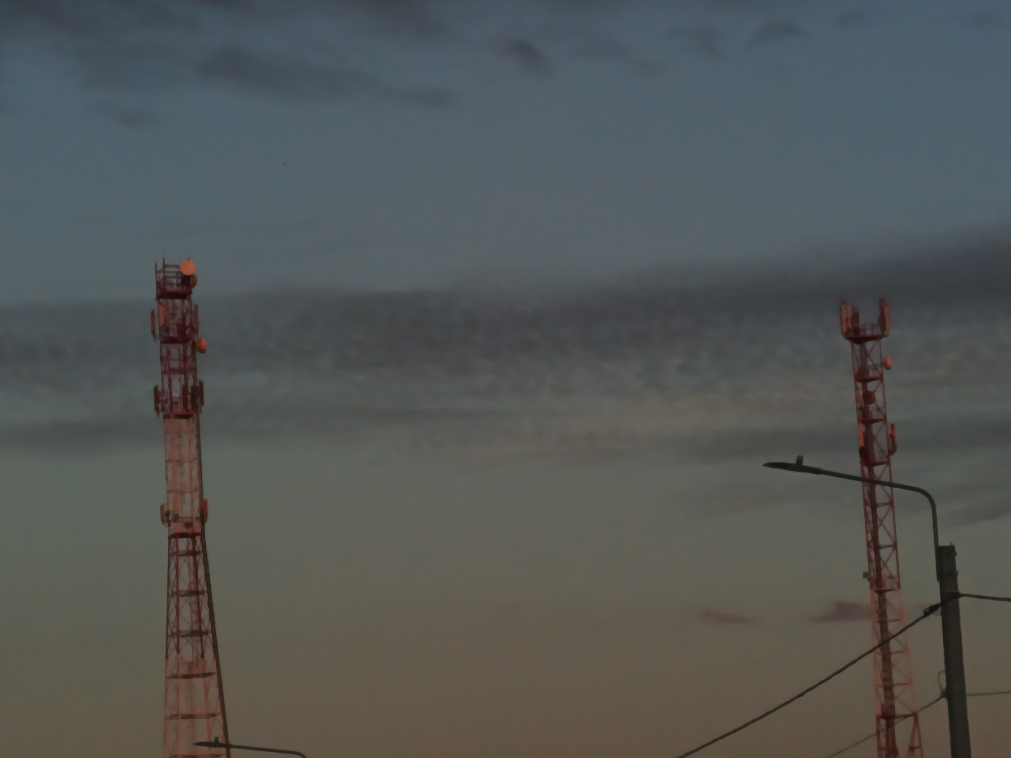 Two cellular towers are visible against a dusky sky with wispy clouds. The tower on the left is taller and more prominent, while the tower on the right is partially obscured by a utility pole and a street light. The towers are constructed of red lattice metal and appear to be functional, with various antennas and equipment attached. The sky transitions from a pale blue at the top to a warmer, orange-pink hue near the horizon, suggesting it is either dawn or dusk. The overall scene is industrial and urban, characteristic of infrastructure found in populated areas. The image does not contain any people or discernible activities.