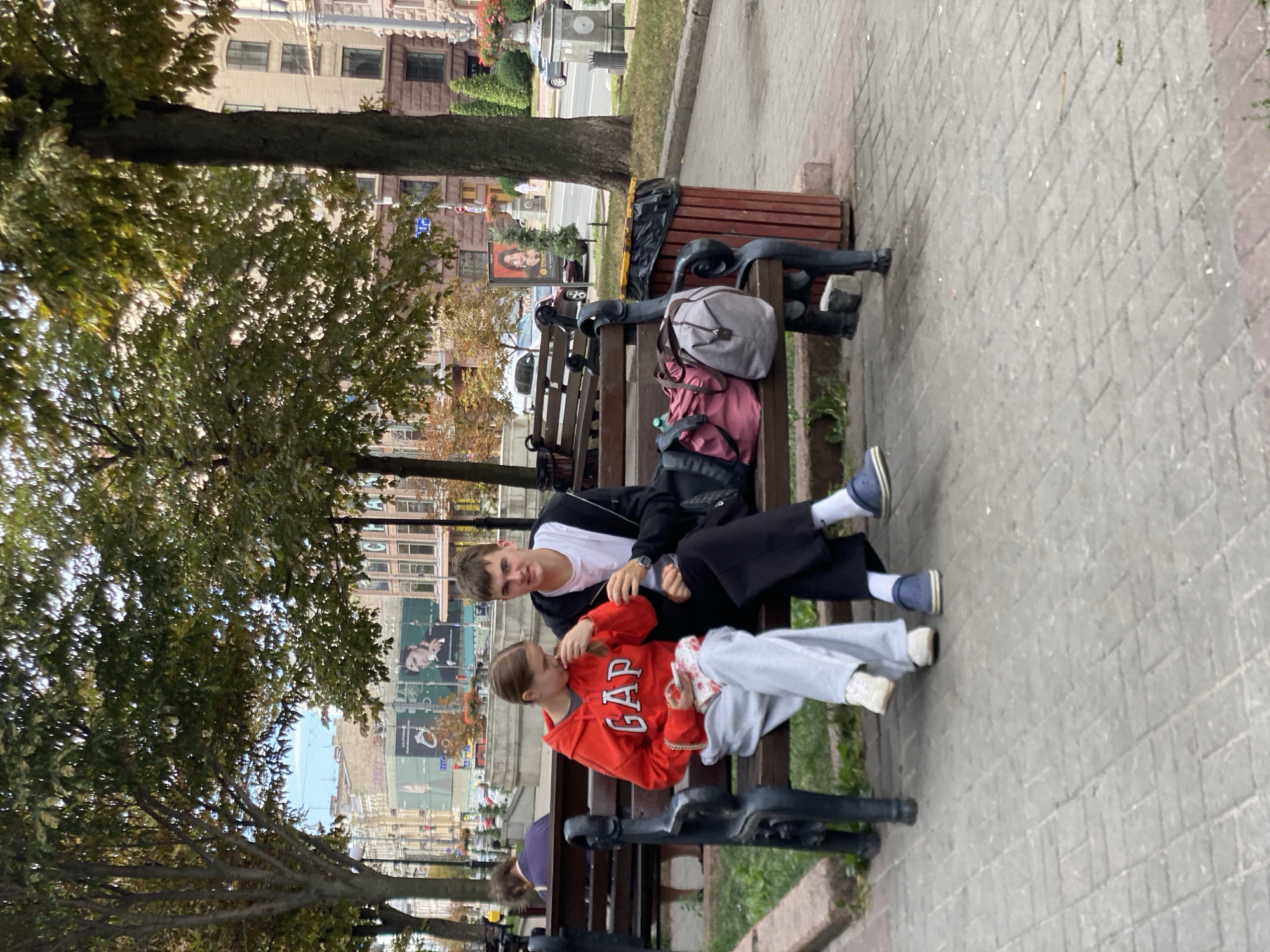 A medium shot shows two young people, a girl and a boy, sitting on a park bench. The girl, on the left, wears a red GAP hoodie, gray sweatpants, and white sneakers. She holds a small pink and white patterned bag and a smartphone. The boy, on the right, is dressed in a white t-shirt, black zipped hoodie, black pants, and blue Crocs. They are seated close together, with the girl looking down at her phone and the boy looking towards the camera.
Several bags are placed on the bench next to the boy, including a pink duffel bag and a gray backpack. A red trash can with a black liner stands beside the bench. The background reveals a city street with buildings, trees, and cars. The ground is paved with light-colored bricks. In the background, a billboard displays an advertisement featuring a woman. The scene appears to be in a public urban park.