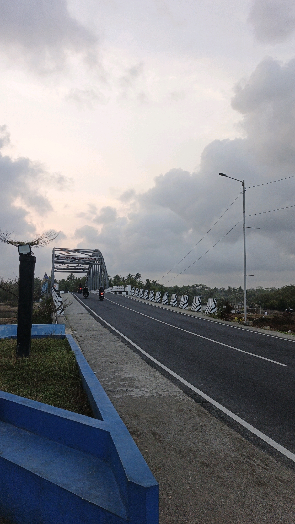 This is a medium shot, captured from a slightly elevated angle, of a road leading onto a bridge in Pangandaran, Indonesia. The sky is overcast with heavy, grey clouds, suggesting either dawn or dusk. The road is asphalt with white lane markings and is flanked by a concrete sidewalk and a decorative blue barrier. On the left side of the road, a black lamp post with a minimalist design stands near a patch of grass.
The bridge itself is a metal arch bridge with a gray, industrial appearance. The text "WIRADINATA RANGGAJIPANG" is visible on the bridge structure. Several motorcycles are proceeding across the bridge, indicating moderate traffic. The sides of the bridge are lined with alternating black and white striped barriers. In the background, a dense line of palm trees and other tropical vegetation is visible, hinting at a coastal or tropical environment. The overall impression is one of a functional infrastructure in a natural, possibly rural, setting.
