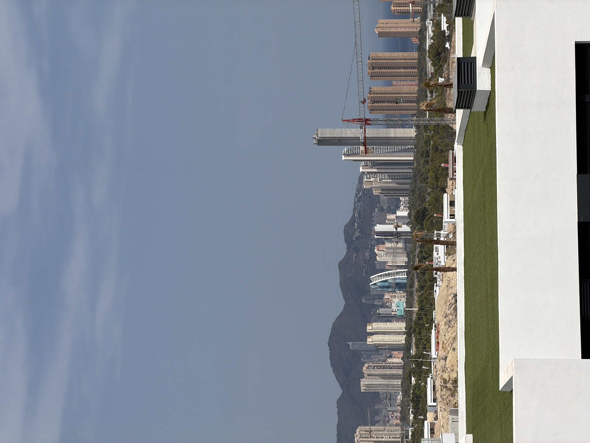 The image shows a panoramic view of the city of Finestrat, Spain, with a foreground of a rooftop terrace and a background of a dense urban skyline and distant mountains. The rooftop terrace features white railings, artificial grass, and ventilation units. In the middle ground, there is a construction site with a prominent red crane. The urban skyline is dominated by numerous high-rise buildings of varying architectural styles and colors, including a distinctive curved skyscraper. Beyond the city, rugged mountains form a natural backdrop under a clear, pale blue sky. The scene appears to be a clear day with good visibility, suggesting it was taken in the daytime. There are no people visible in the image.