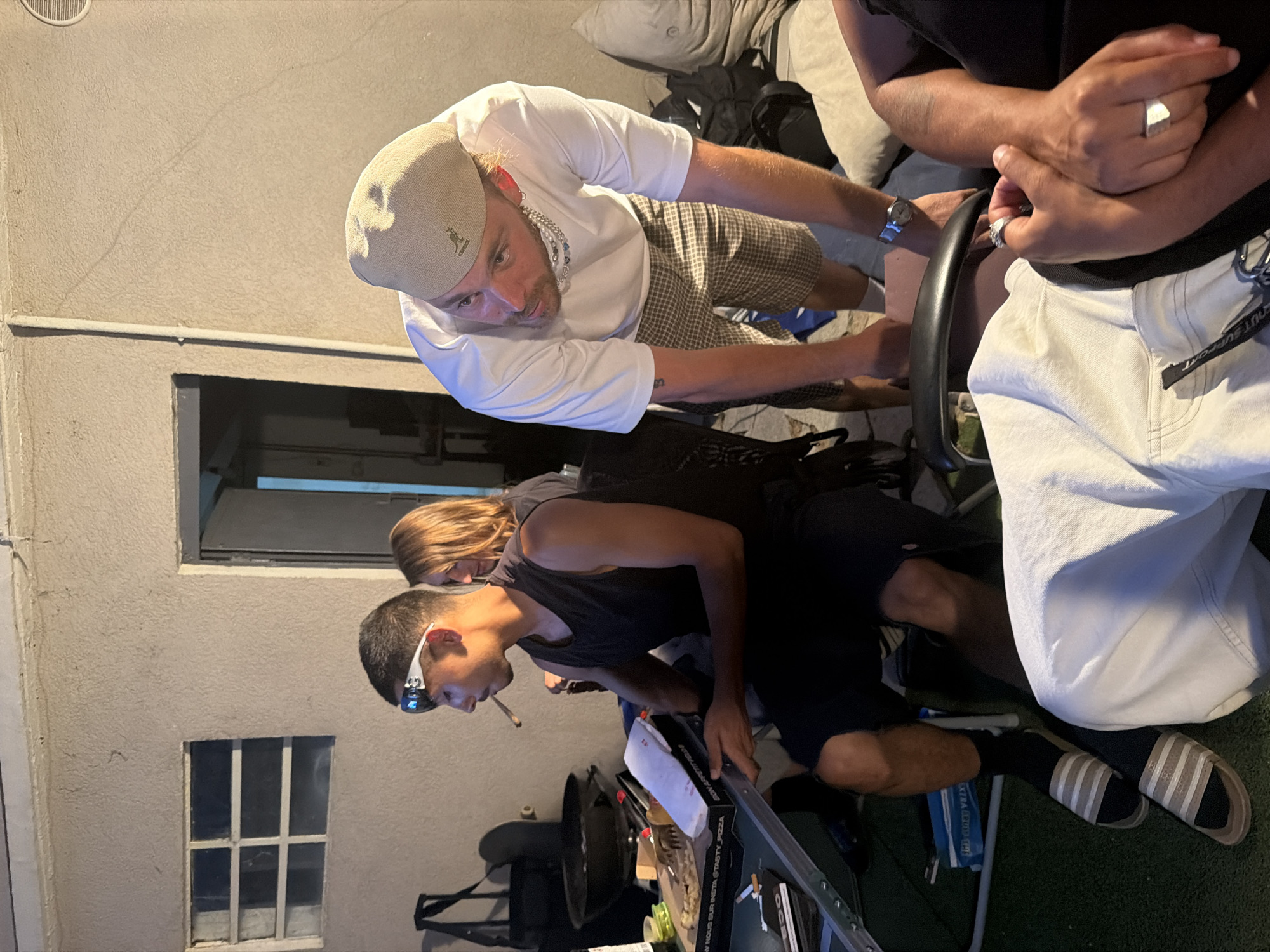 The image depicts an outdoor scene, possibly a backyard or patio, at dusk. In the foreground, a person wearing white pants and a black shirt is seated, with their hands clasped. To the left, a man in a black tank top and dark shorts is sitting at a metal table, wearing white sports glasses and smoking a cigarette. A pizza box and other items are on the table. A woman with blonde hair is standing behind him. To the right, a man wearing a beige Kangol hat, a white t-shirt, and plaid shorts is leaning over the table, looking towards the seated individuals. The background shows a building with a window and a doorway. There are chairs, bags, and other miscellaneous objects scattered around the area. The lighting suggests artificial illumination and natural twilight. The city of Villeurbanne, France is not explicitly identifiable from the visual cues within the image.