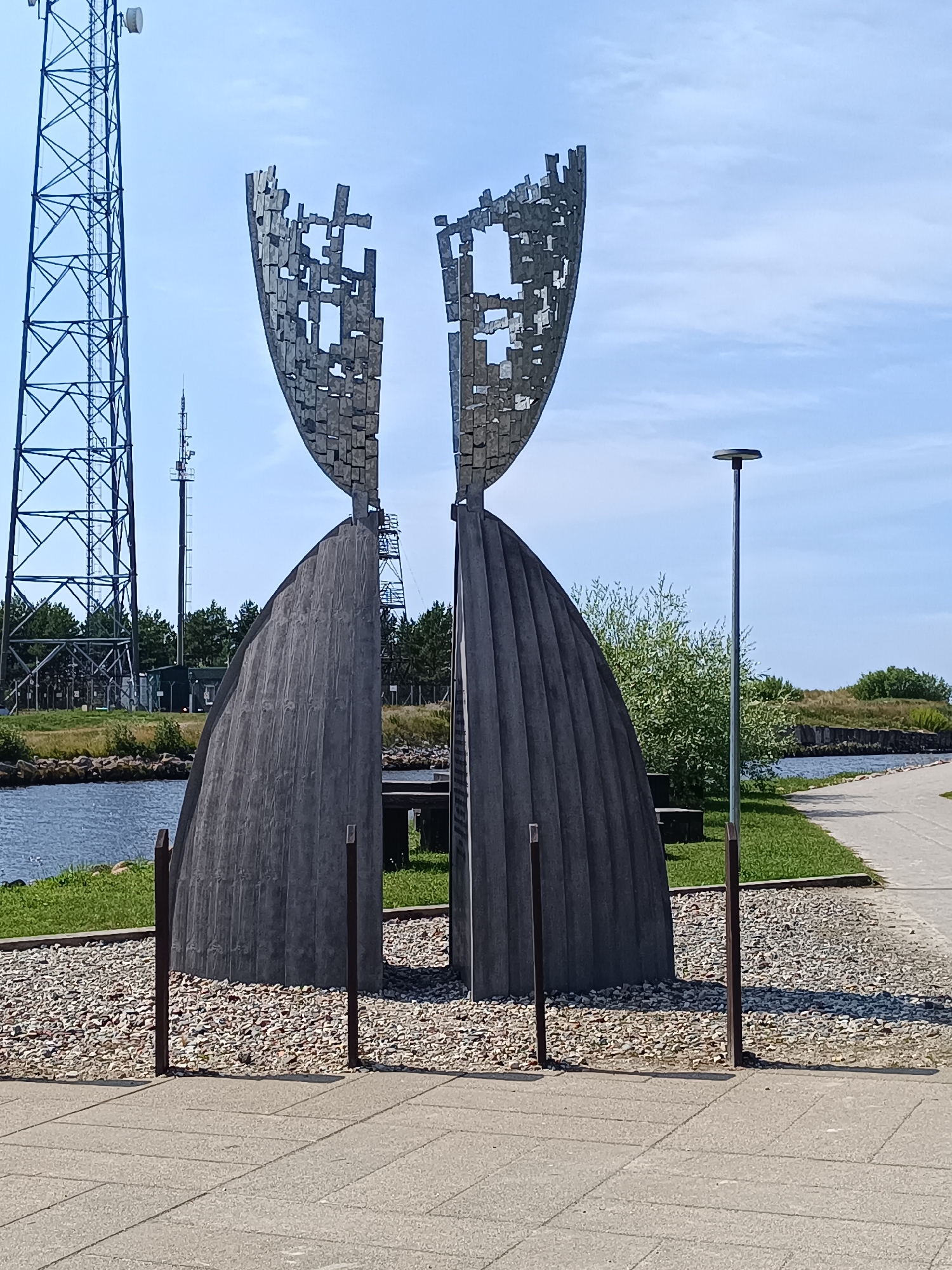 The image shows a public monument in Pāvilosta, Latvia. The monument is composed of two large, curved, dark gray, fluted concrete structures that meet at the top, creating a central gap. Above this, two abstract metal sculptures, resembling shields with cut-out geometric patterns, are positioned to form a mirrored pair, further widening the gap. The monument stands on a bed of light-colored gravel, demarcated by short, dark metal posts. In the foreground, a paved walkway made of light gray concrete slabs extends across the frame. Behind the monument, a body of water, likely a river or sea, is visible, with a grassy embankment and trees on the far side. To the left of the monument, a tall, lattice metal telecommunications tower with an antenna on top is prominent against a clear blue sky with scattered clouds. Another smaller telecommunications tower is visible behind the monument. A single, slender lamppost stands to the right of the monument. The overall setting appears to be a waterfront area, possibly a promenade or park. There are no people visible in the image.