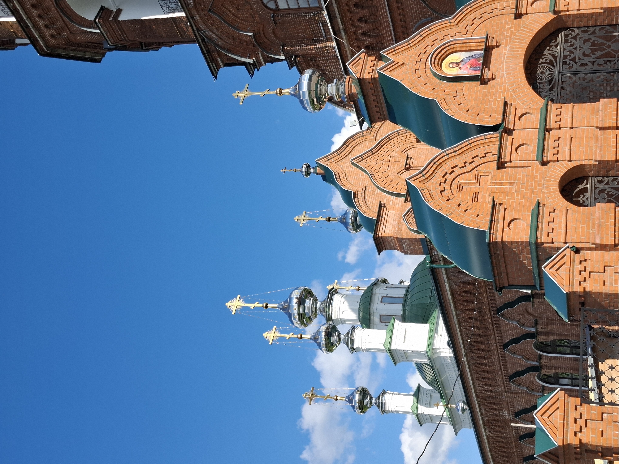 This is a low-angle, full-shot photograph of a brick Russian Orthodox church against a bright blue sky with scattered white clouds. The church features multiple onion domes topped with golden crosses. The domes are of varying sizes, with some being silver and others white. The building is constructed of red brick, with decorative arches and trim in white. A green roof is visible on a lower section of the church, and a metal fence with an ornate gate is in the foreground. A small icon of Jesus Christ is mounted above the gate. The architecture suggests it is the Annunciation Cathedral in Kungur, Russia, known for its unique red brick construction and numerous domes. There are no people or animals visible in the image. The scene appears to be bathed in sunlight, indicating daytime.