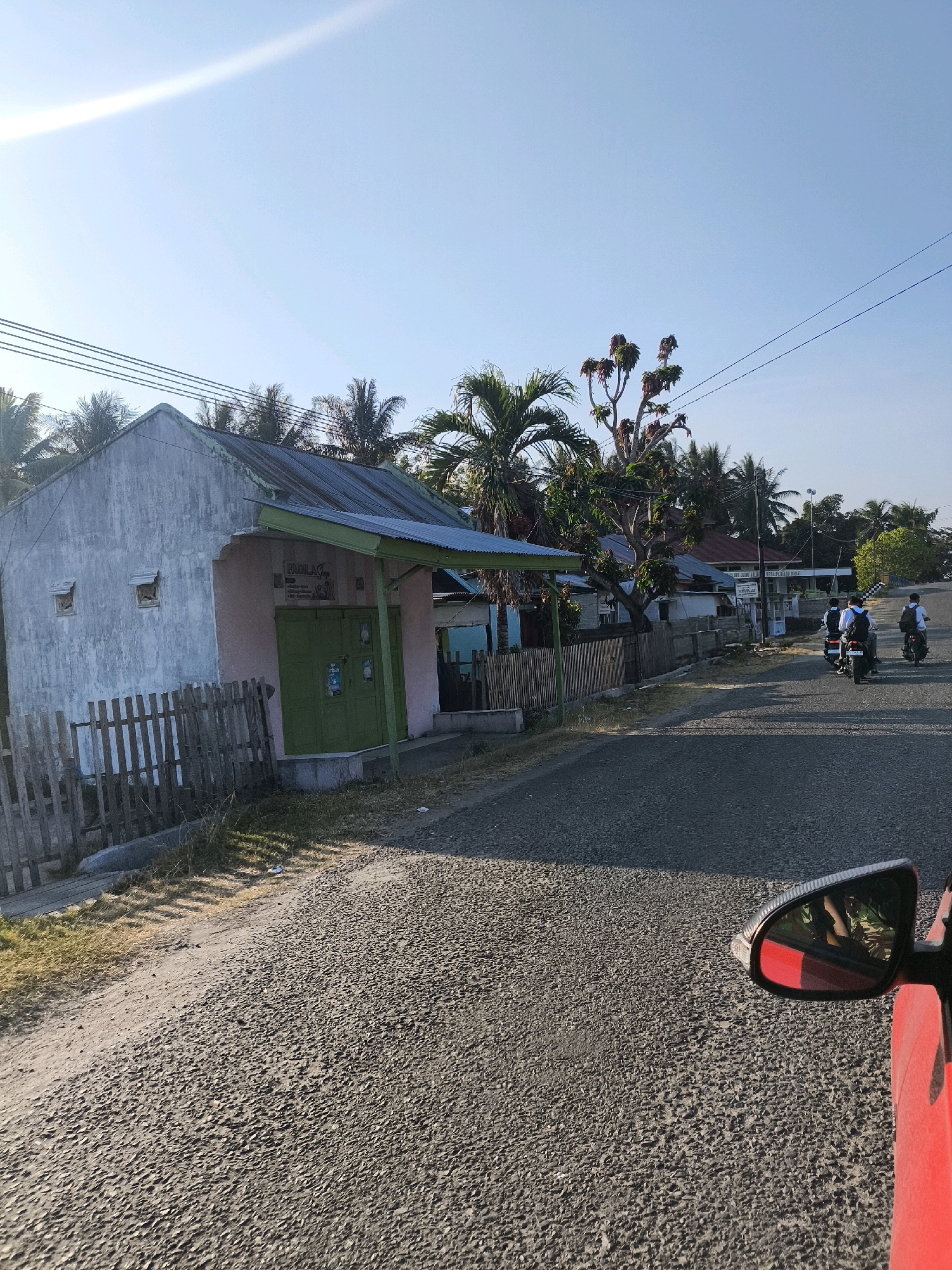 The image depicts a sunlit street scene in Tilamuta, Indonesia. A weathered, pale pink building with a green door and corrugated metal roof is situated on the left, with a wooden fence in front of it. The building appears to be a small shop or residence. Across the street, several people on motorcycles are visible, some wearing backpacks, suggesting they might be students or commuters. The environment is characterized by palm trees, other vegetation, and a clear blue sky. The road surface is asphalt, and the lighting indicates it is daytime, likely morning or afternoon. The perspective suggests the photo was taken from inside a vehicle, with a red car mirror visible in the foreground.