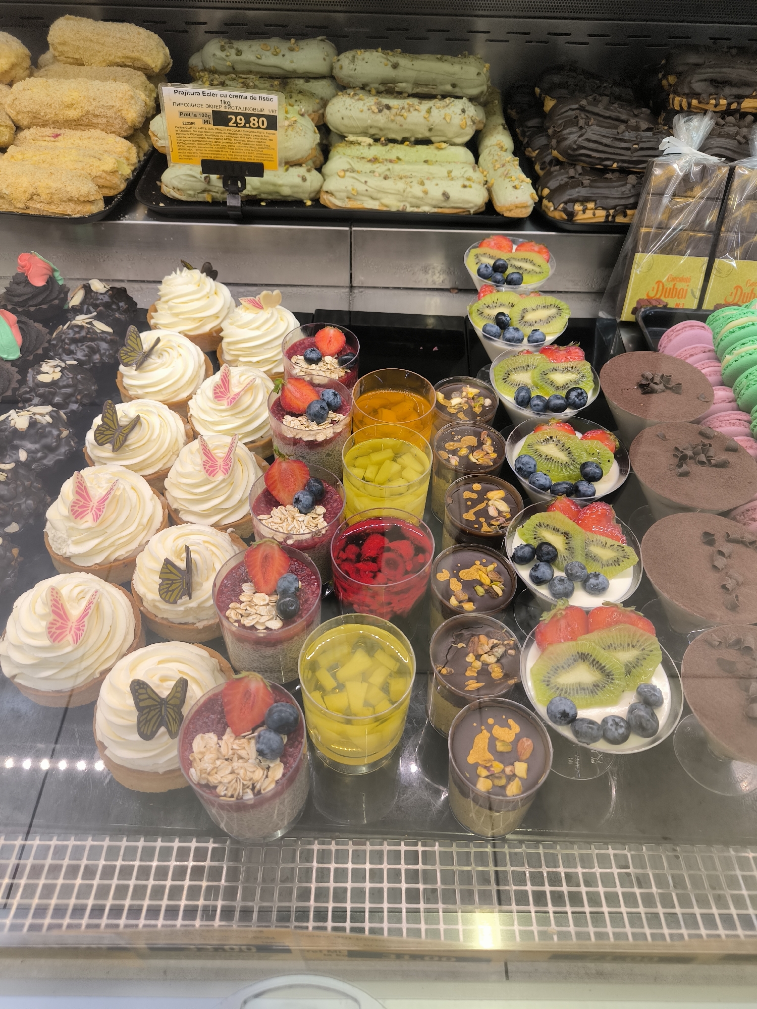 The image shows a display case of various pastries and desserts, likely in a bakery or supermarket in Chișinău, Moldova. In the foreground, there are several small dessert cups filled with layered ingredients, topped with fruit, granola, and nuts. To the left, there are white frosted cupcakes decorated with edible butterflies. Behind these, there are dark chocolate cupcakes. In the middle row, there are more white frosted cupcakes and what appear to be chocolate eclairs with a dark glaze and toppings. Further back, there are lighter colored eclairs, some with a pale green glaze and pistachios, and others with a yellow coating. On the right side of the display, there are rows of macarons in pink and green, and larger chocolate desserts, possibly mousse or tarts, dusted with cocoa powder. A price tag is visible for "Prajitura Ecler cu crema de fistic," indicating a price of 29.80. The overall environment is a refrigerated display case, suggesting a commercial food setting. There are no people visible in the image.