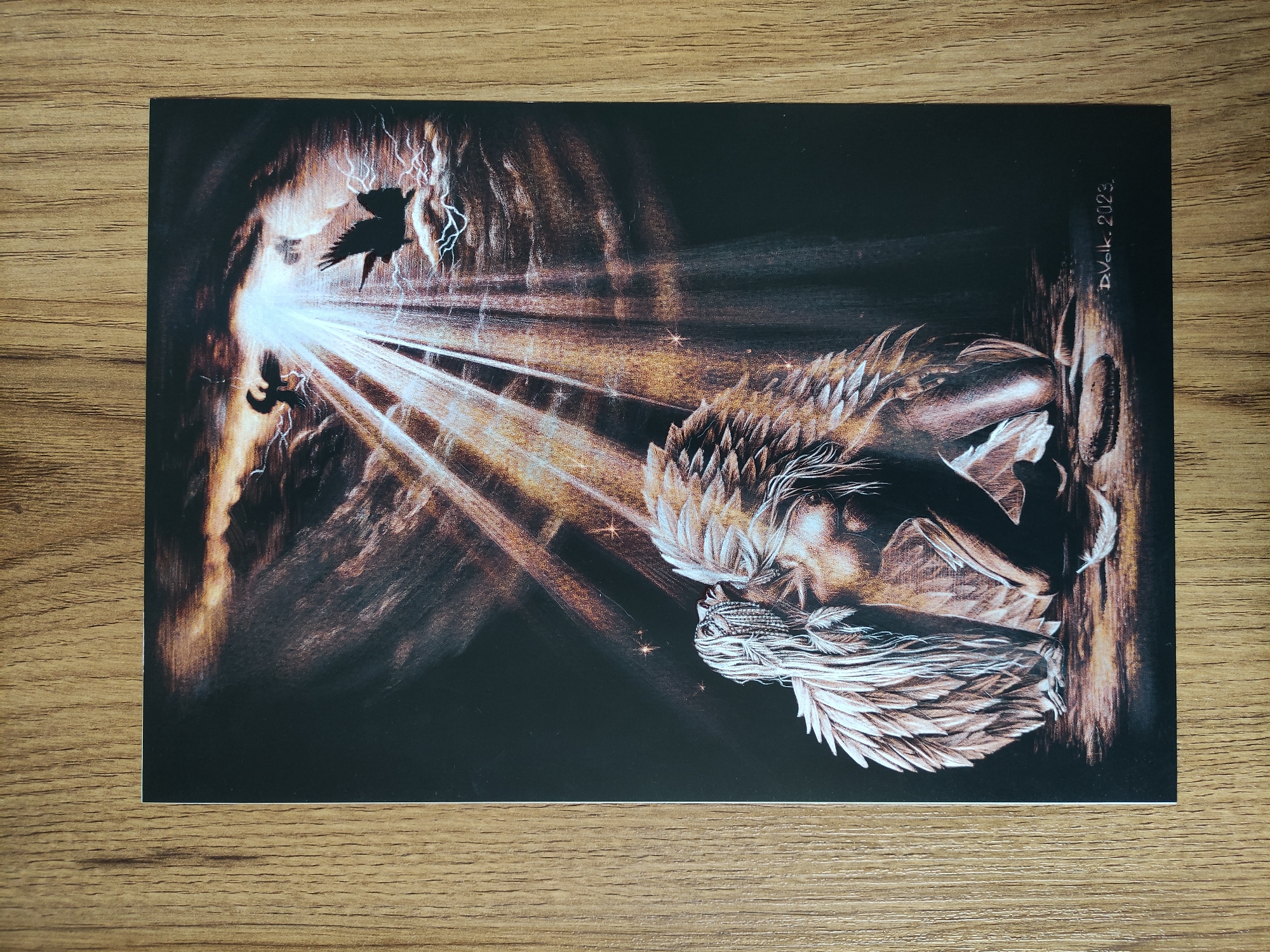 The image is a print or artwork on a dark background, placed on a wooden surface. It depicts an angel-like figure with feathered wings and hair, seemingly kneeling or crouching. Light rays emanate from a bright area to the left, where two bird-like silhouettes are visible. The background appears to be a dark, cavernous space with lightning-like streaks. "D. Volk. 2023." is written in the upper right corner. The location is not identifiable as Tbilisi, Georgia.