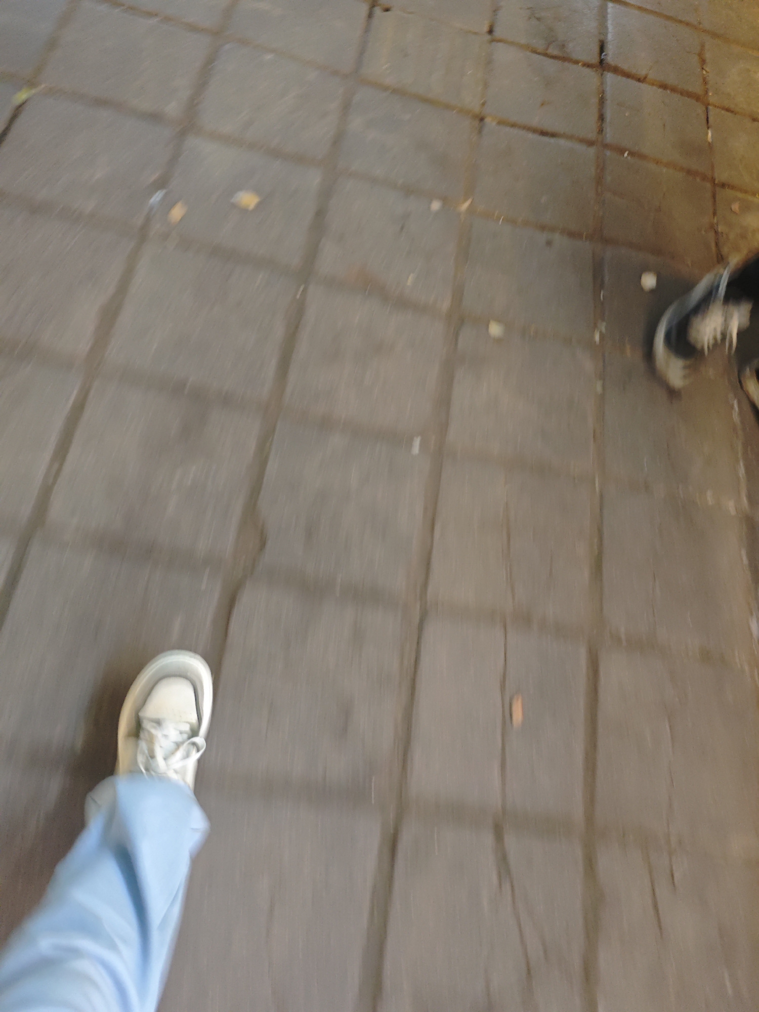 The image shows a first-person view of someone walking on a paved surface, likely a sidewalk or pedestrian area in the Shapingba District, China. The ground is made of square stone or concrete pavers. The person is wearing light blue pants and white sneakers. Another person wearing a black/gray shoe is visible to the right.