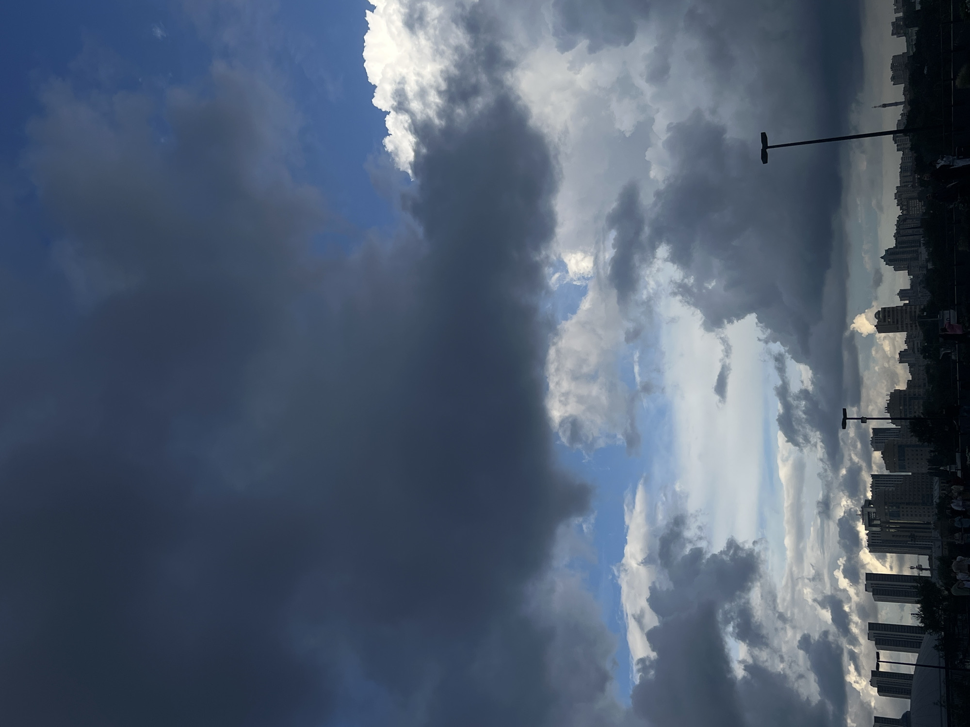 The image depicts a panoramic view of the Yekaterinburg city skyline under a dramatic sky filled with a mix of dark, ominous storm clouds and patches of bright blue sky with fluffy white clouds. In the foreground, silhouetted against the sky, are several tall, modern buildings and skyscrapers, characteristic of a city's urban landscape. Several streetlights are visible, including one prominently placed in the mid-right of the frame. Below the skyline, at ground level, a few individuals are present, appearing as small, indistinct figures, suggesting they are observing the sky or the cityscape. The overall atmosphere is one of impending weather change, with the contrast between the dark clouds and the clear sky creating a visually striking scene.