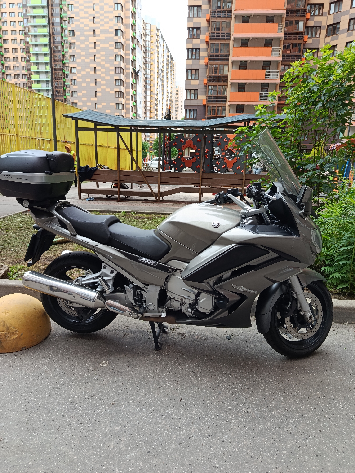 The image depicts a grey Yamaha FJR1300 motorcycle parked on an asphalt surface. The motorcycle has a black top case mounted on the rear. In the background, there are tall, modern apartment buildings with a mix of beige, orange, and green facades. A yellow fence runs along the left side of the image, bordering a covered wooden structure that appears to be part of a playground or recreational area. Green trees and bushes are visible on the right side of the frame. A person wearing a yellow jacket is standing near the covered structure. The scene suggests an urban or suburban residential area. The lighting indicates it is daytime, and the sky is overcast, suggesting cloudy weather. No specific activities are clearly depicted, other than the motorcycle being parked and the person standing. The overall impression is a still, everyday scene.