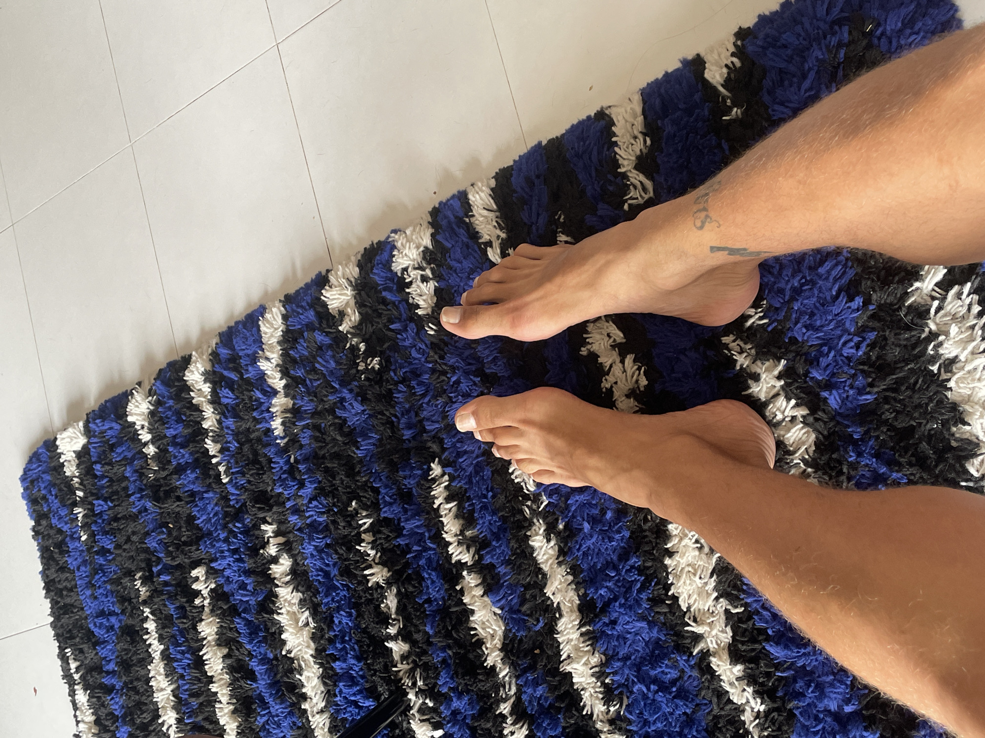A pair of legs and feet are positioned on a shaggy rug with alternating stripes of white, blue, and black. The rug lies partially on a tiled floor. The legs are fair-skinned, and the lower leg has a tattoo. The environment appears to be indoors. There is no information to support the location being in Chang Phueak, Thailand.
