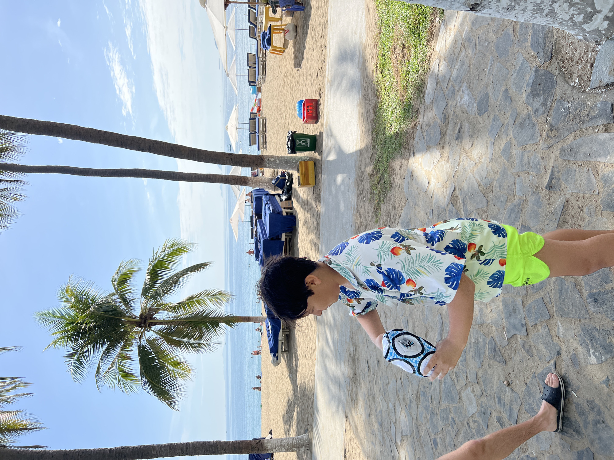 The image depicts a young boy holding a football on a paved walkway at a beach resort in Nha Trang, Vietnam. The boy is wearing a floral-patterned short-sleeved shirt and bright green shorts. In the background, a sandy beach is visible with rows of lounge chairs and white umbrellas. The ocean stretches out beyond the beach, meeting the horizon under a clear blue sky with scattered clouds. Palm trees frame the scene, with two tall palm trees in the foreground and a larger one behind them. The walkway is paved with irregular grey stones, and on the beach, several trash bins are placed near the lounge areas. There are a few people scattered on the beach and in the water. The scene suggests a relaxed, tropical vacation atmosphere.
