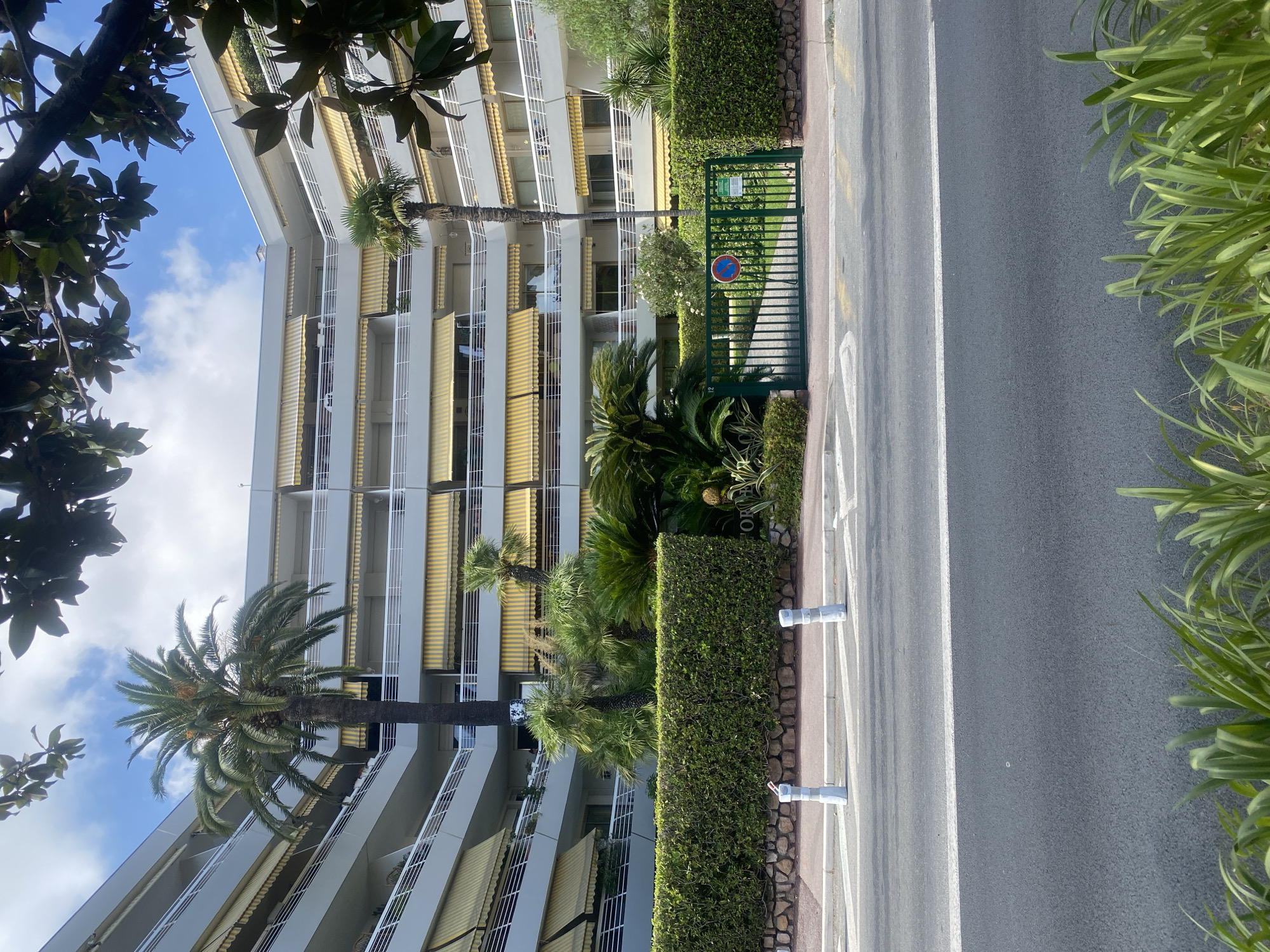 The image depicts the exterior of a multi-story apartment building with a curved facade, situated in Antibes, France. The building features white balconies with gray railings and numerous yellow awnings. Palm trees and lush green hedges are visible in front of the building. A green metal gate with a "no parking" sign is present, leading to a driveway. The scene is viewed from a street, with a road and sidewalk in the foreground. The sky is partly cloudy.