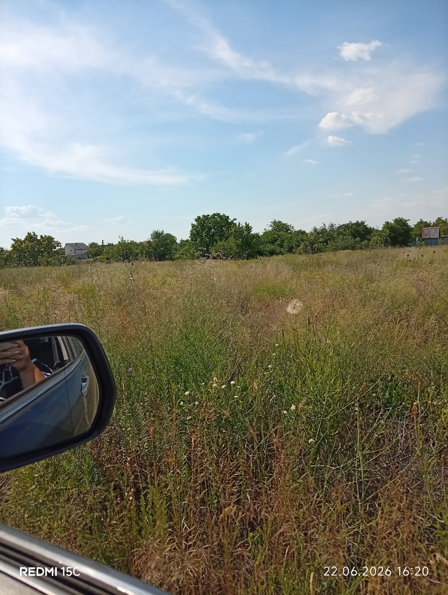 The image depicts a rural landscape viewed from inside a vehicle, with the side mirror prominently in the foreground on the left. Through the mirror, a person's hand is visible, holding what appears to be a smartphone. The main scene shows a dry, grassy field extending towards a line of trees and houses in the distance. The sky is blue with scattered white clouds. The date stamp "22.06.2026 16:20" and "REDMI 15C" are superimposed on the bottom of the image. The environment suggests a warm, sunny day in a rural area. The presence of houses and trees indicates a settlement or village.