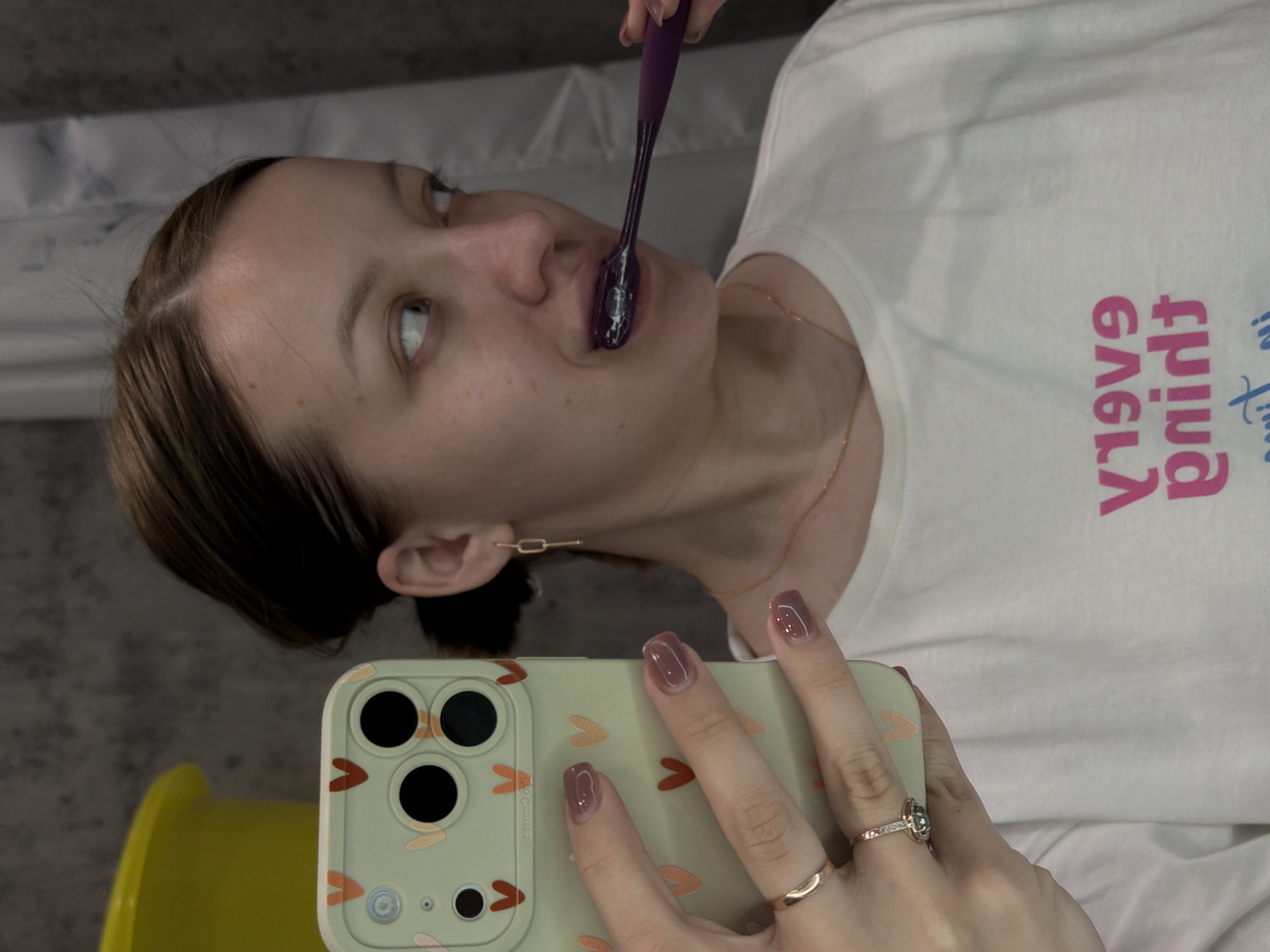 A woman is brushing her teeth with a purple toothbrush while taking a selfie with a smartphone in a light green case decorated with orange hearts. She is wearing a white t-shirt with pink lettering, a gold necklace, and gold earrings. She has mauve-colored nail polish and is wearing a gold ring on her ring finger. A yellow object is partially visible in the lower left corner of the frame. The background appears to be a bathroom setting with a white surface. There is no indication of the location being Reshetnikovo, Russia.