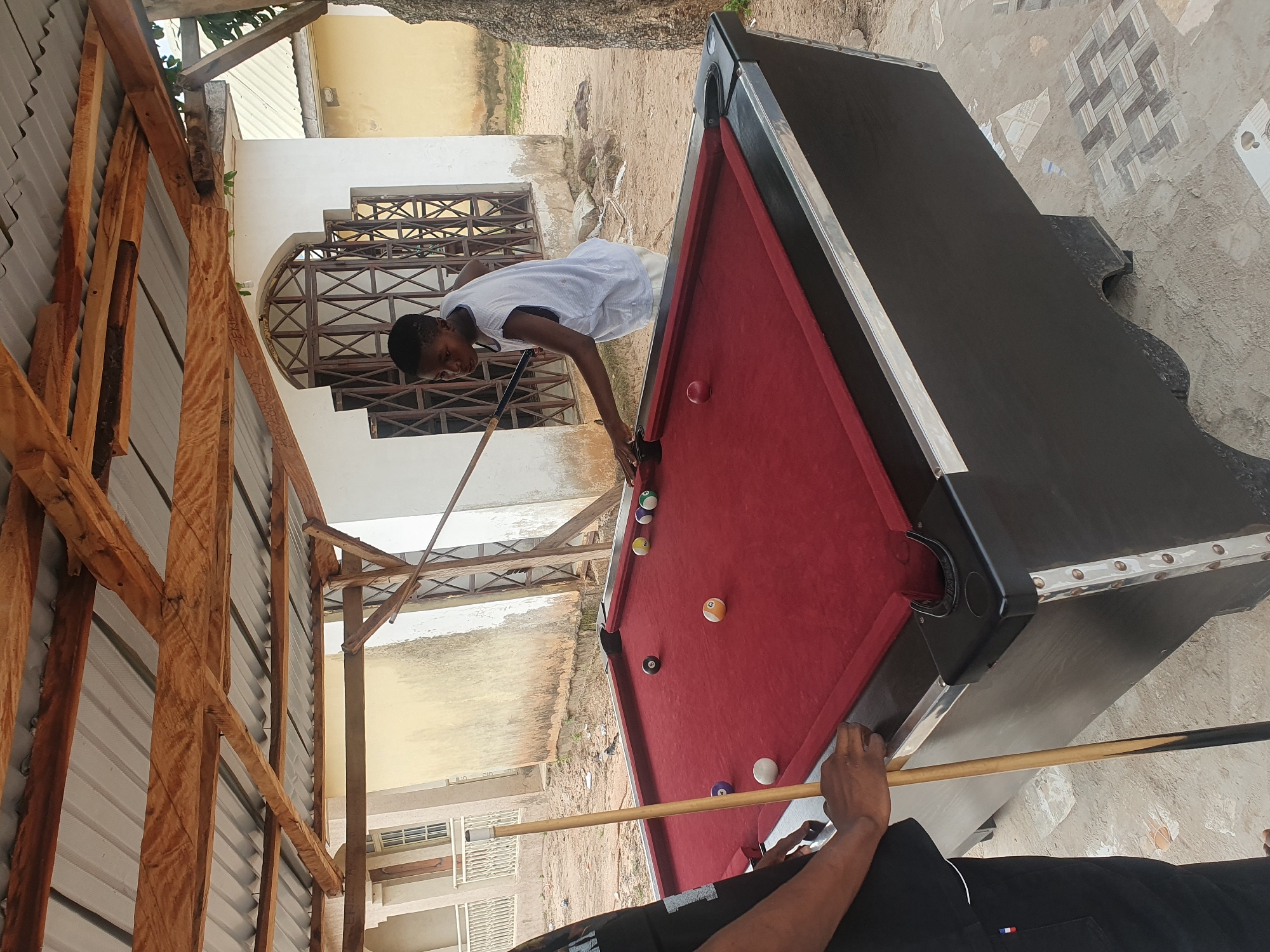 In Lokoja, Nigeria, an outdoor pool table covered with a red felt surface is situated beneath a rustic, wooden-framed roof. The table is a standard size and appears to be in fair condition, with black sides and silver trim. Multiple billiard balls are scattered across the table, including solid colors and stripes. A young male, wearing a light blue sleeveless shirt and light-colored shorts, is actively engaged in playing pool. He is positioned at one end of the table, holding a pool cue and appearing to take a shot. Another individual, partially visible on the left side of the image and wearing a black t-shirt and black pants, also holds a pool cue, suggesting they are also participating in the game. The background features a white building with arched windows and a metal grate, indicating a somewhat weathered and possibly rural or less developed setting. The ground around the table is sandy with some scattered debris and decorative tiles near the table's base. The overall scene depicts a casual game of pool taking place in an open-air environment in Nigeria.
