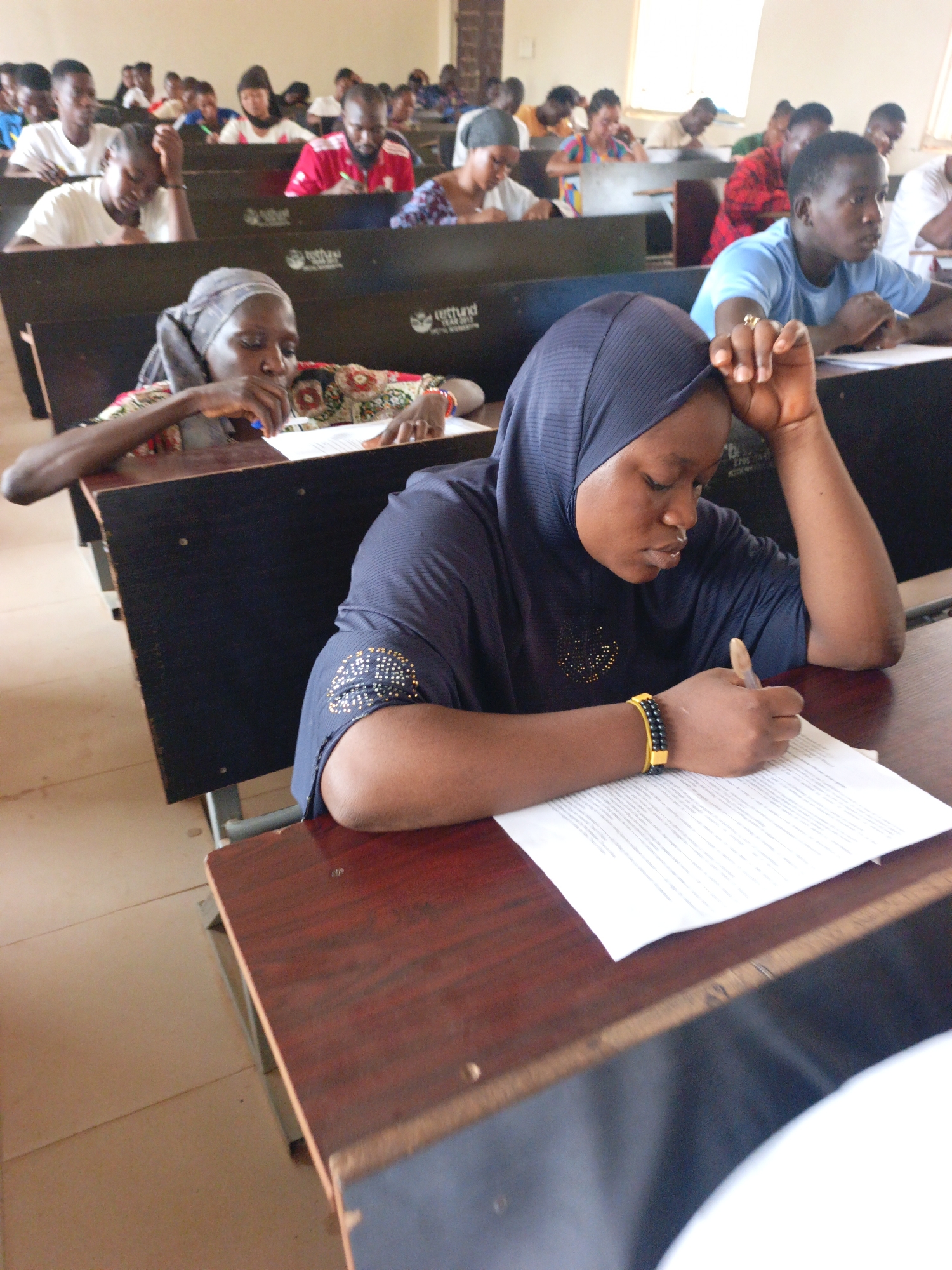 The image depicts a classroom setting filled with students engaged in writing on paper. The students are sitting at desks with dark panels. One student in the foreground wears a dark blue hijab and a yellow and black bracelet, focused on writing. Another student to her left wears a headscarf with a patterned shirt. Several other students are visible in the background, filling the rows of desks. The room is lit by daylight coming through a window, with pale beige walls. The specific city and location are unknown.