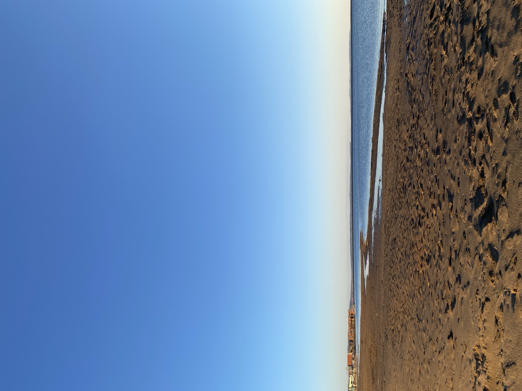 The image depicts a beach scene in Chaikovsky, Russia, with a clear blue sky dominating the left side of the frame. Adjacent to the sky is a narrow strip of water, possibly a river or bay. To the right of the water is a stretch of dark, wet sand, marked with footprints. In the background, along the horizon, there is a row of buildings.