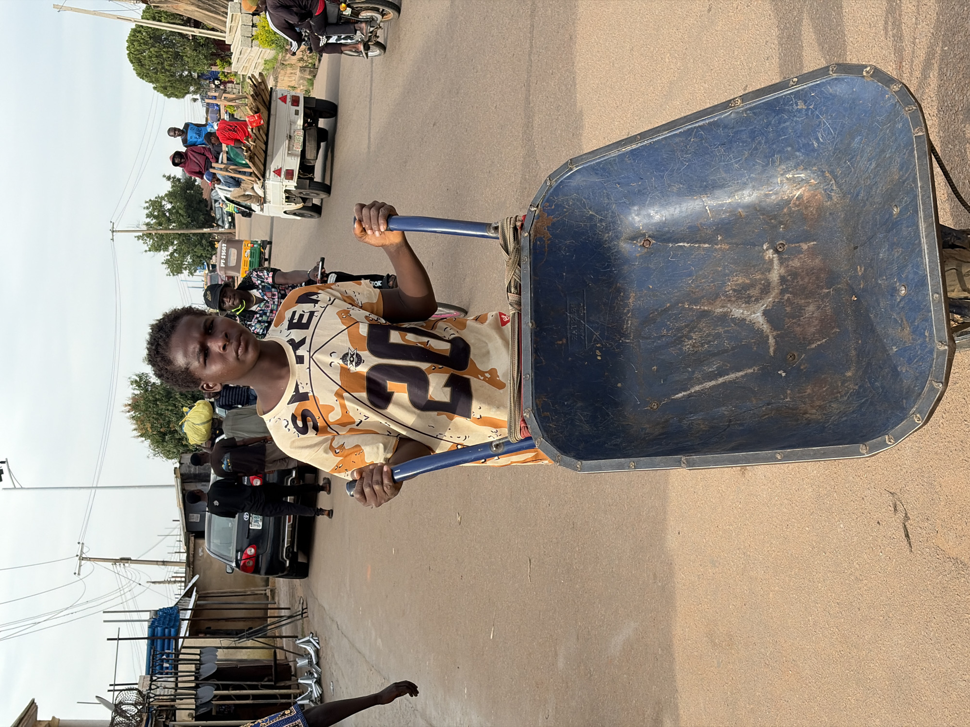 A young person is pushing a blue wheelbarrow down a dusty road in Gura Top, Nigeria. The person, who appears to be wearing a light-colored t-shirt with a graphic design and dark pants, is looking directly at the camera. The wheelbarrow is empty and shows signs of wear and tear. In the background, there are several vehicles, including a black car, a small white truck carrying lumber, and a tricycle. A few other people are visible on the road and sidewalks, some of whom appear to be carrying goods. The environment is urban with buildings, power lines, and trees lining the road. The sky is overcast.
