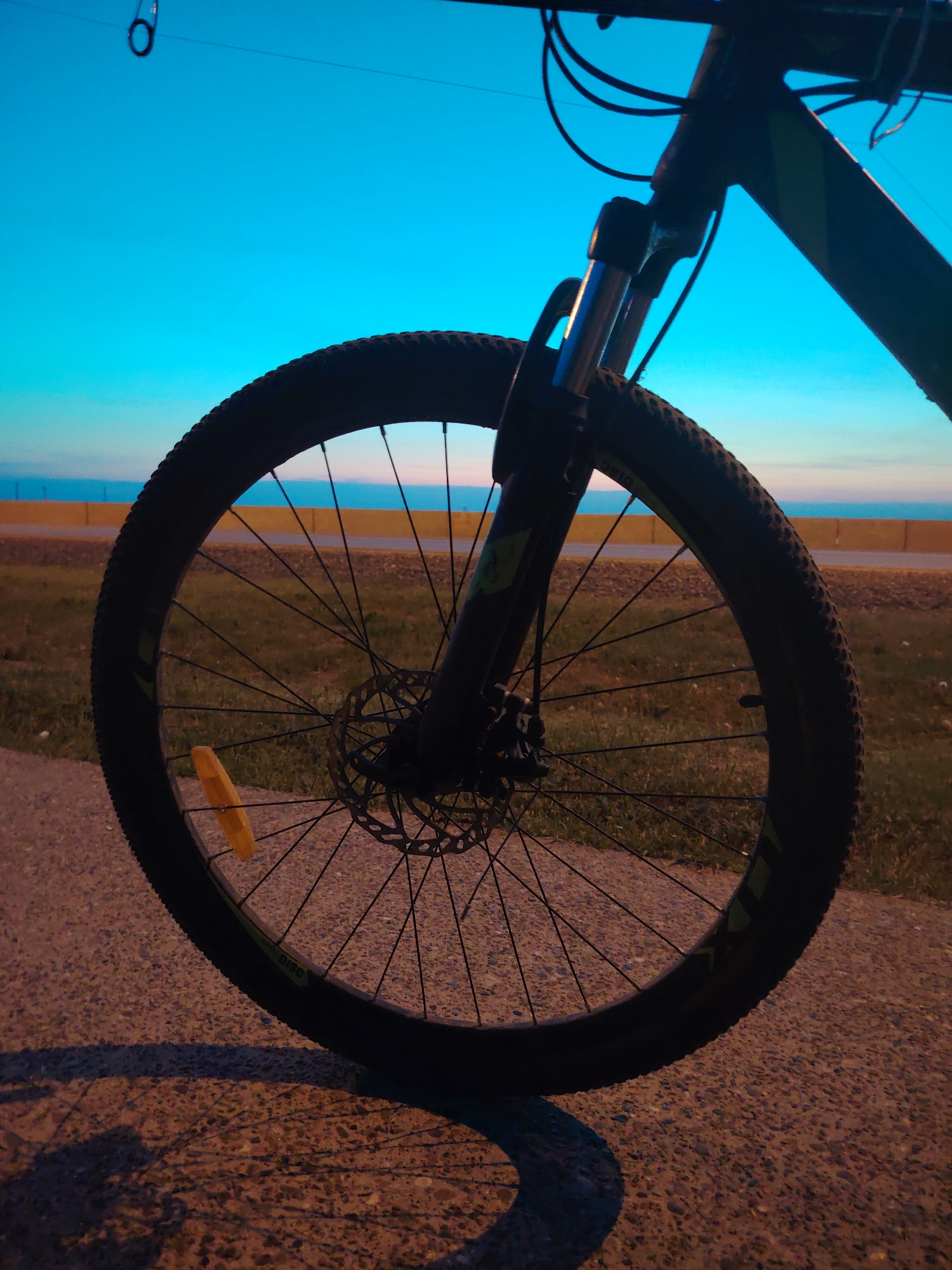 The image shows the front wheel of a mountain bike parked on a paved surface with a view towards the Kama river in Chaikovsky, Russia. The bike has a disc brake and a yellow reflector on the spokes. The background includes a yellow retaining wall, grass, the river, and a light blue sky. The time appears to be around dusk or dawn based on the lighting.