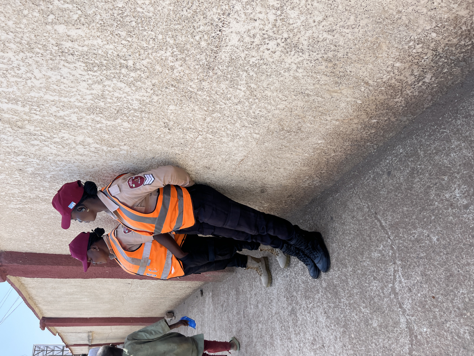 Two uniformed women stand side-by-side outdoors in Jos, Nigeria. Both wear maroon caps, khaki shirts with badges, orange safety vests, and black pants. One wears black boots, the other beige. A concrete wall is behind them. Other people are partially visible in the foreground.
