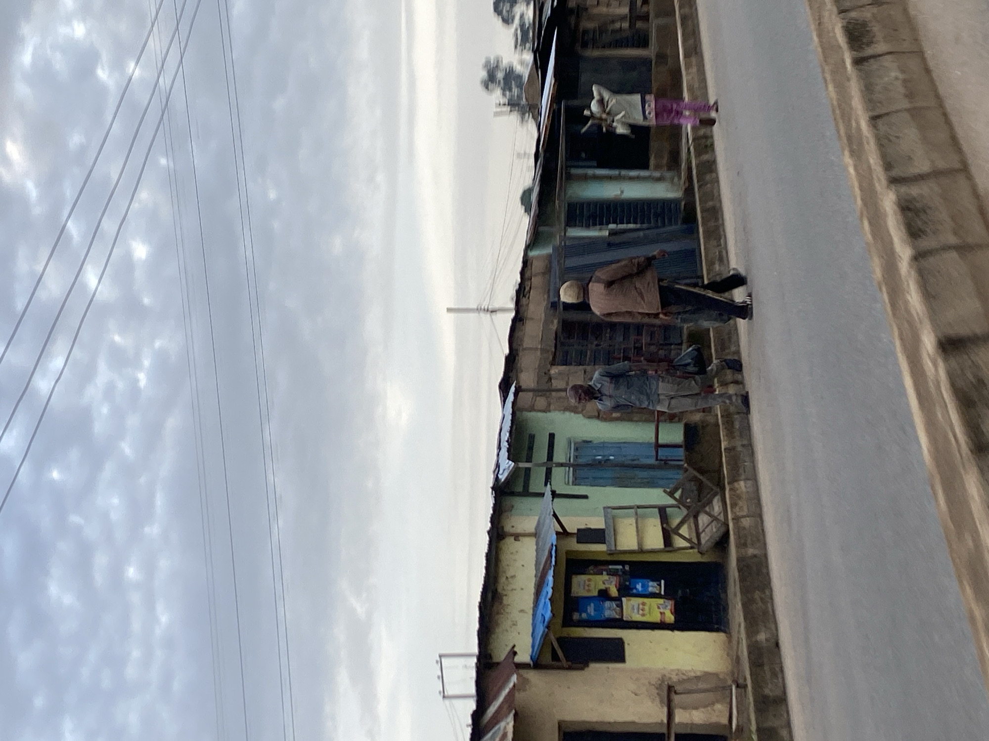 The image depicts a street scene in Jos, Nigeria, under an overcast sky. In the foreground, a paved road runs parallel to a row of small, weathered buildings. These structures appear to be shops or market stalls, characterized by corrugated metal roofs, some with tin and others with what looks like a blue tarp. The buildings are painted in various muted colors, including yellow, green, and beige, with visible wear and tear.
Three individuals are walking along the sidewalk in front of the shops. The person on the left is a man in a grey long-sleeved shirt and dark pants. He is carrying a black bag over his left shoulder. In the middle, a man in a brown jacket and dark pants walks with a tool or stick in his right hand. To the right, a person in a light-colored jacket and pink patterned pants walks away from the camera, carrying an item over their right shoulder.
Several objects are visible near the shops, including a shelving unit outside a yellow building displaying posters, and what appears to be a collapsed wooden structure or cart. Electrical wires are strung across the upper portion of the image, connecting to poles in the background. The overall impression is of a humble, bustling urban environment with signs of everyday commerce and life.