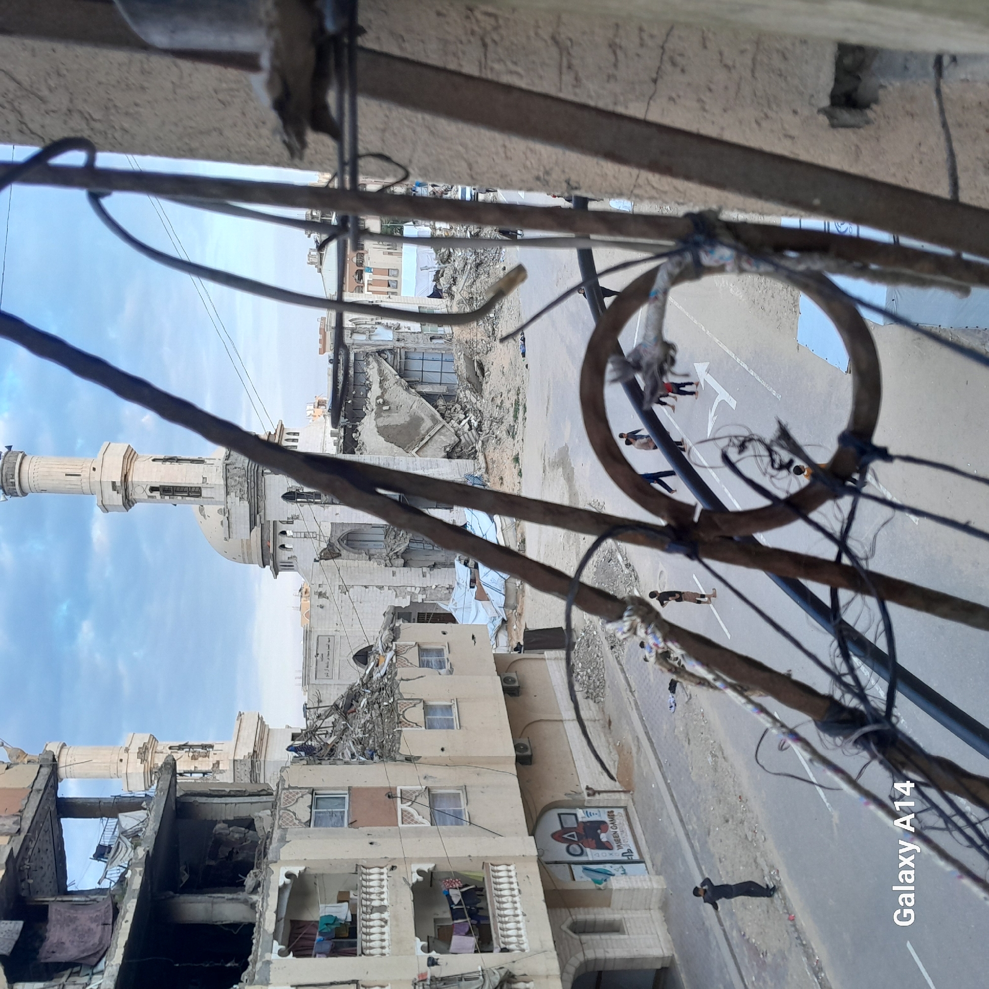 This image, taken in Khan Yunis, Palestinian Territory, depicts a street scene viewed from an elevated position, likely a window or balcony, framed by blurry, rusty metal bars and tangled wires in the foreground. The sky is overcast and light blue.
On the left, a multi-story light-colored building, possibly residential, displays significant structural damage with exposed interiors, hanging laundry on balconies, and numerous small impact marks on its facade. The ground floor features an arched entrance with "YASEEN GAMES" text, a PlayStation logo, and console button symbols (Δ, O, X, □).
In the background, a large, light-colored mosque stands with two minarets and a central dome, all exhibiting damage, including visible cracks and missing sections. An arched entrance with Arabic script is visible at its base, surrounded by debris. To the right of the mosque, buildings are reduced to extensive piles of rubble and partially collapsed structures, with white tarpaulins visible among the debris.
The asphalt street in the midground features white double solid lines and a right-turn arrow marking. Debris and dirt line the road edges. Two dark bundles of fabric or material are visible on the left side of the street near the curb. Approximately 5-7 individuals, appearing to be mostly children or young adults, are scattered on the right side of the street, some barefoot, standing or walking. On the left side of the street, one individual in dark clothing stands near the damaged building, facing right.