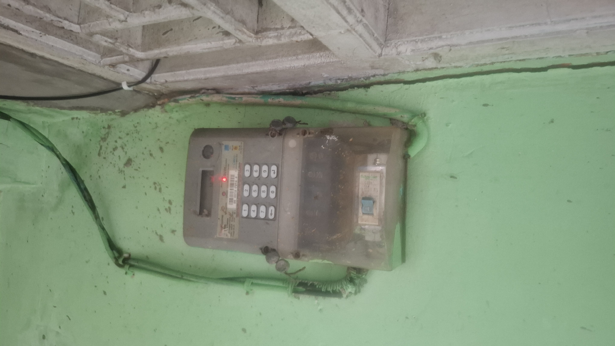 An outdoor electrical meter is mounted on a pale green wall. The meter is gray and rectangular, with a clear plastic cover revealing internal components. A keypad with numerical buttons and an arrow is visible on the upper part of the meter, along with a red light. Two small, dark gray padlocks are attached to the sides of the meter. Green electrical conduit runs vertically behind and to the left of the meter, connected by a twist tie. To the right of the meter, a white barred structure is visible, partially obscuring a concrete wall. The wall appears weathered and stained in places. The scene is illuminated by natural light.