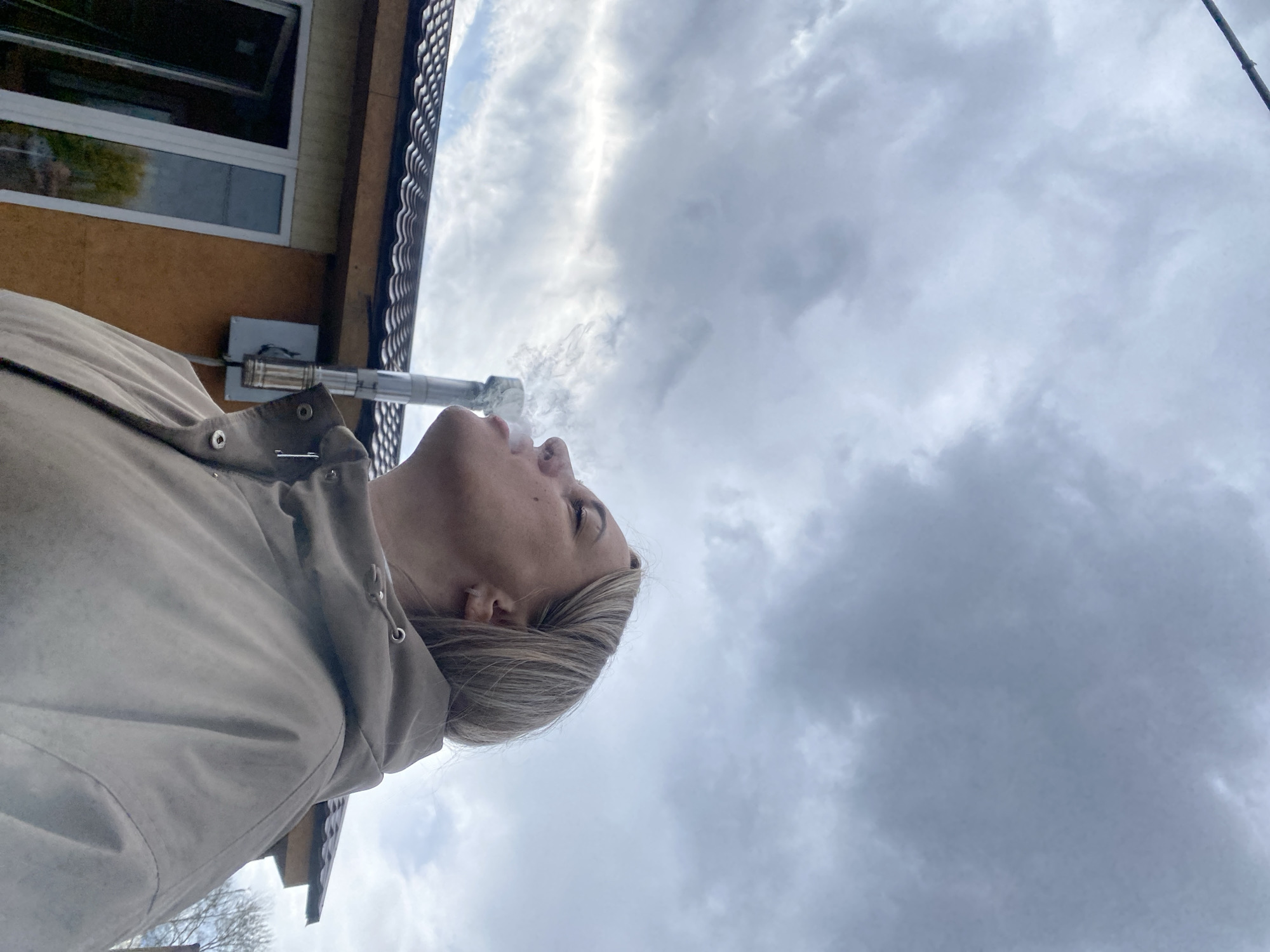 The photograph, taken outdoors, features a woman smoking a device, possibly a vape or e-cigarette. She is positioned with her head tilted back, looking towards the sky. The smoke she is exhaling is visible against the backdrop of the cloudy sky. She is wearing a light-colored jacket with a hood. To the left of the woman, there is a building with a window visible. There is also a metallic pipe, likely part of the building's ventilation system. The environment suggests an overcast day. The location details of "Unknown, Unknown" are not helpful in determining an actual location.