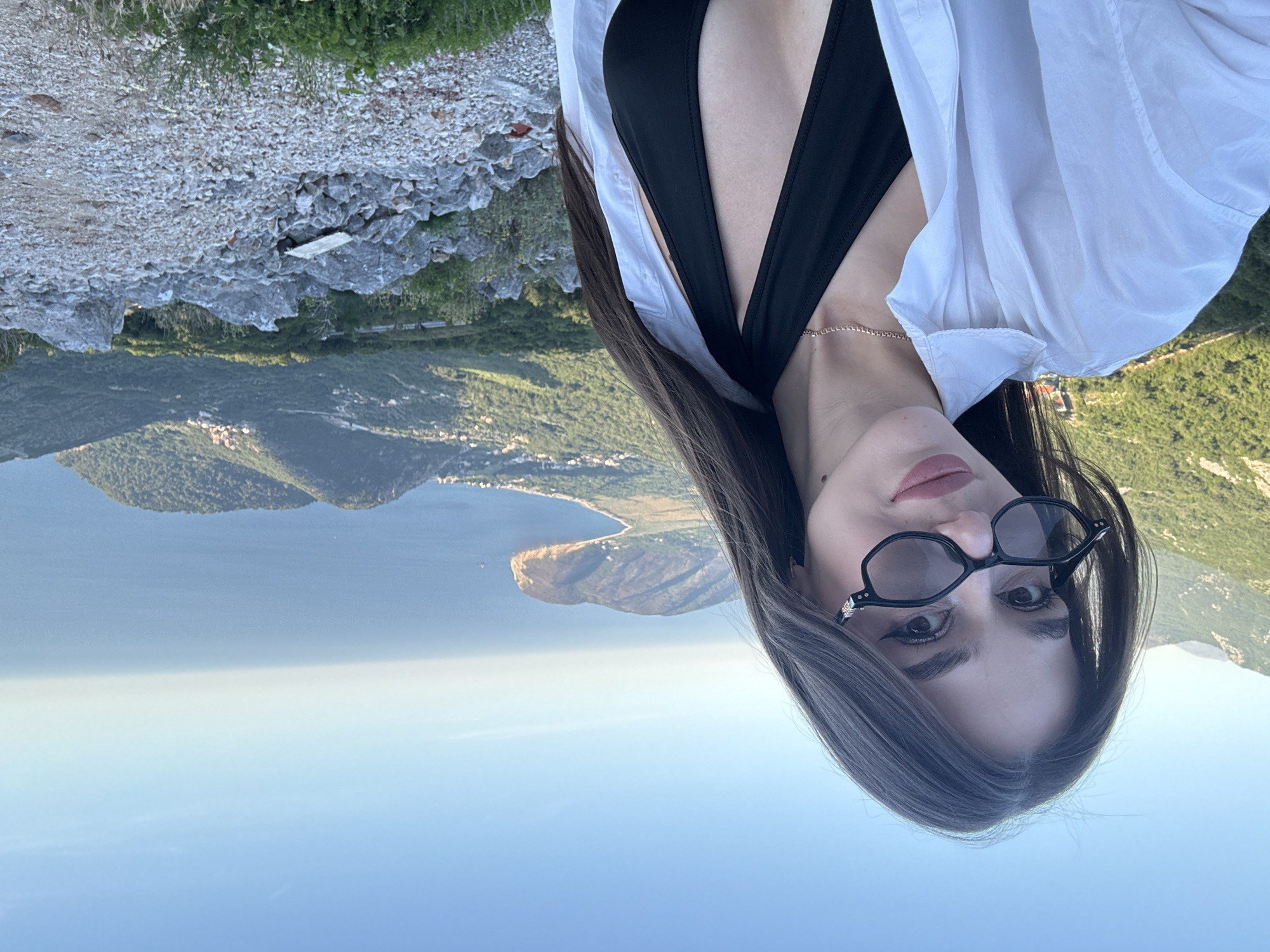 A woman with dark hair and glasses poses in Novoselje, Montenegro. She is wearing a black top and a white shirt. The background includes the Adriatic Sea, mountains, and the coastline of Montenegro. The scene also includes rocky terrain.