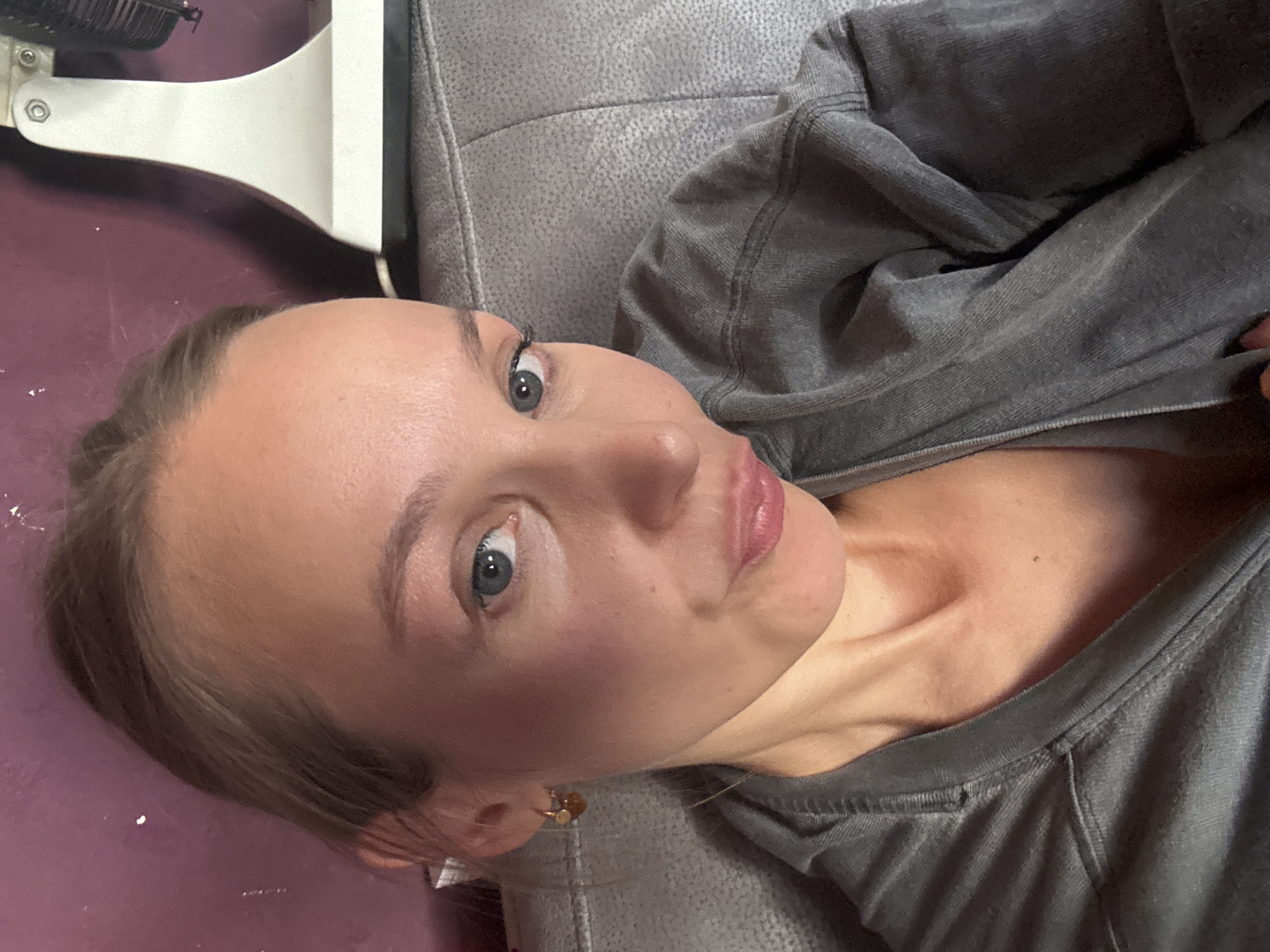 This is a close-up, selfie-style photograph of a young woman with blonde hair pulled back. She is wearing a grey hooded sweatshirt. Her head is tilted slightly, and her expression is playful, with her lips pursed. She has blue eyes and is wearing a small, gold hoop earring with a charm. The background shows a purple wall and a portion of a white object that appears to be a piece of furniture or an appliance. The woman is sitting on a grey, textured surface, likely a sofa or chair. The image provides no information about the location in Überherrn, Germany.
