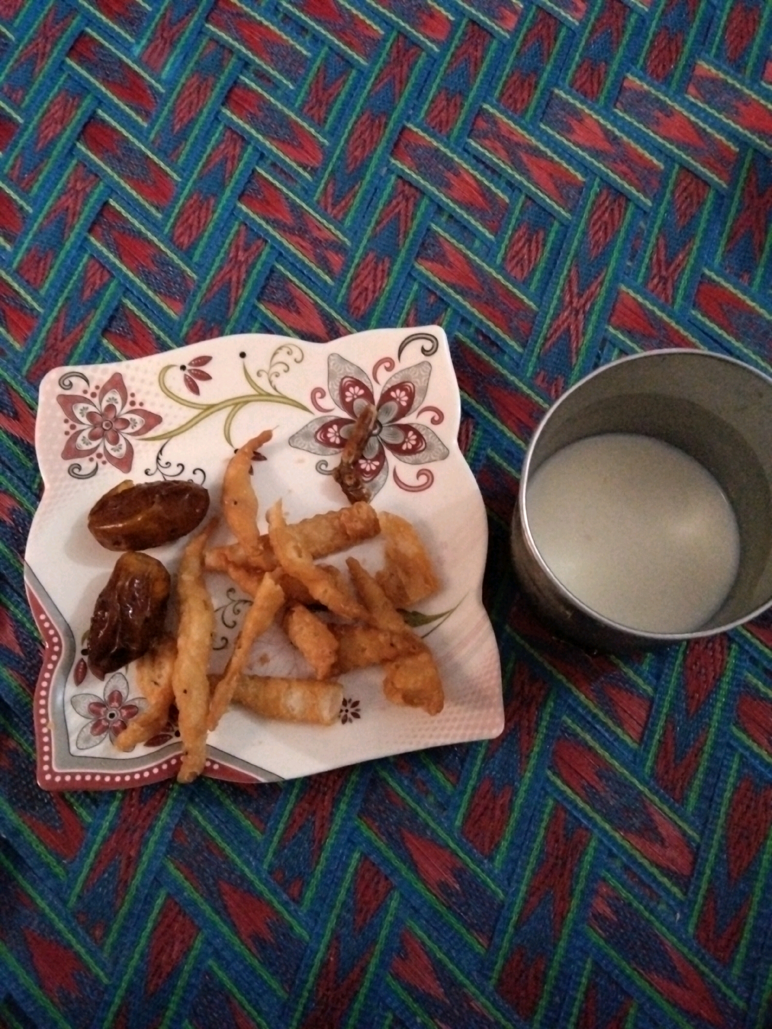 A plate of dates and fried food sits on a woven mat. To the right of the plate is a metal cup filled with a pale liquid, likely milk. The mat has a red, blue, and green woven pattern. The scene appears to be an indoor setting, possibly a home in Shahbaz Khel, Pakistan, given the cultural context suggested by the food items and the traditional mat. There are no people visible in the image. The lighting is dim, suggesting it could be evening or an overcast day.