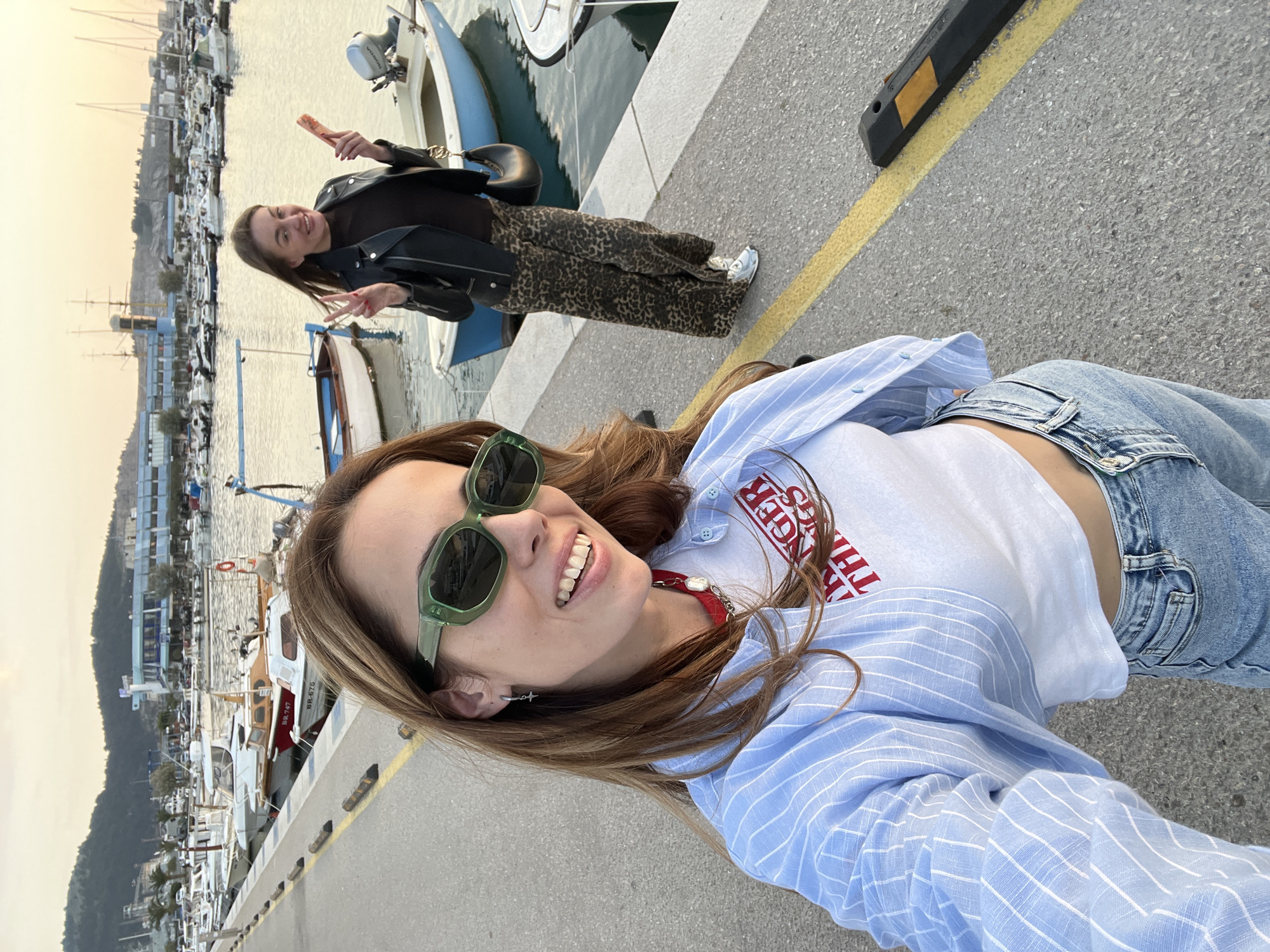 The image captures two women in a harbor setting. The woman in the foreground is smiling at the camera, wearing light-wash denim jeans, a white cropped t-shirt, and a light blue striped button-up shirt. She is also wearing green sunglasses. In the background, another woman is standing near a boat, holding a food item and making a peace sign. She is wearing a black leather jacket and patterned pants. The location is a harbor in the city of Bar, Montenegro, as evidenced by the presence of boats and the coastal landscape visible in the background. A yellow line is painted on the concrete ground.