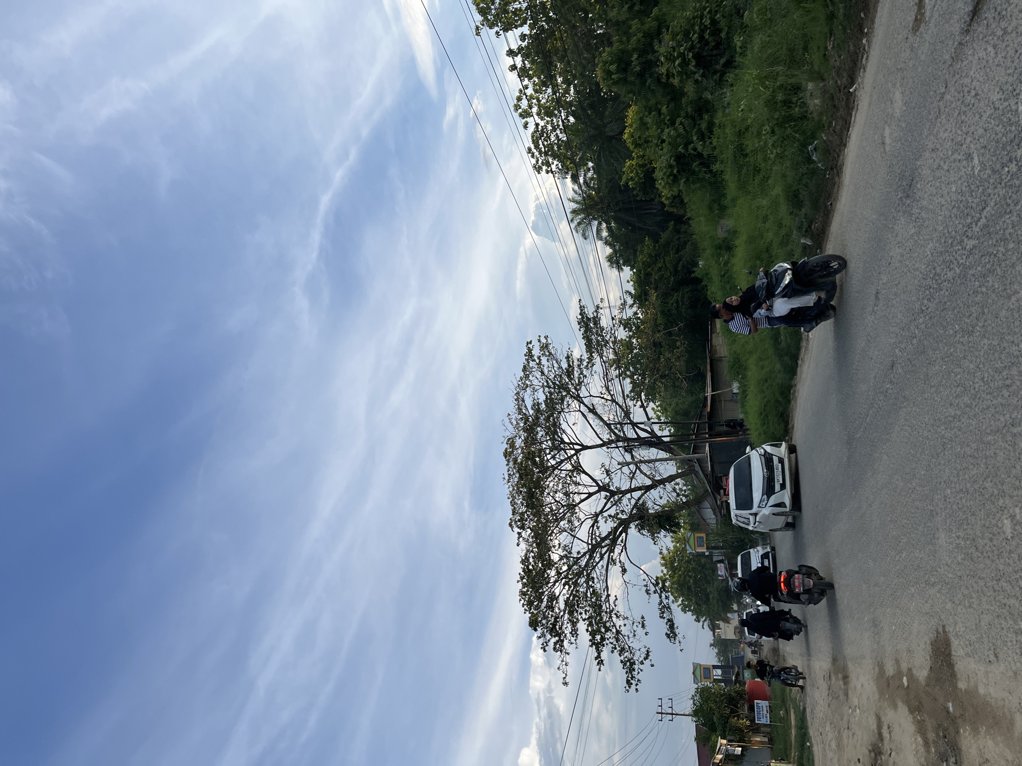 This media file captures a scene on a paved road in Benuaraja, Indonesia, with a bright blue sky and scattered wispy clouds suggesting it's daytime. The road curves slightly, bordered by lush green vegetation on the left and a line of trees on the right.
Several vehicles are present on the road. The most prominent are motorcycles, with a few riders visible. One motorcycle in the foreground has two people on it, the rider wearing dark clothing and the passenger in white and a black head covering. Further down the road, other motorcycles are also seen. A white car is parked on the right side of the road, and another car is visible behind it.
A large, bare-branched tree with sparse leaves is a significant feature on the left side of the frame, its branches reaching towards the sky. Power lines are strung across the scene, indicating infrastructure. Some small buildings and signs are visible along the roadside, hinting at a populated area. One sign near the bottom right appears to have text, but it is not clearly legible. The overall atmosphere suggests a moment of travel or daily life unfolding on a somewhat busy road under a pleasant sky.