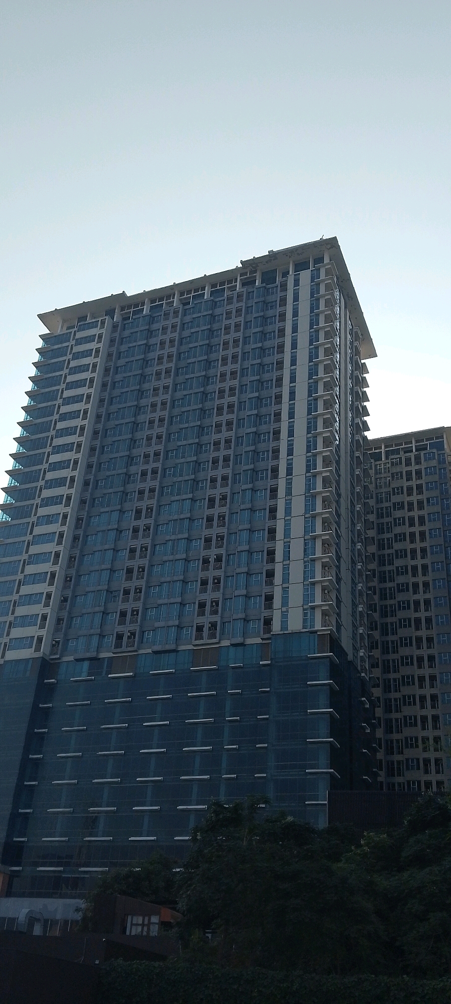 The image shows a close-up, low-angle view of two very tall, modern apartment buildings under construction in Bekasi, Indonesia. The primary building in the foreground is a massive skyscraper with a blue-tinted glass facade covering its upper floors. The lower portion of this building features a different design with horizontal lines and a darker blue hue. The building on the right is partially visible and appears to have a similar architectural style. Both buildings are characterized by numerous windows and balconies. The sky above is clear and a pale blue, suggesting it is daytime. At the bottom of the image, dense, dark green foliage from trees obscures the base of the buildings and the ground level. There are no people visible in the image. The scene depicts an urban construction site, focusing on the imposing scale and modern design of the residential towers.