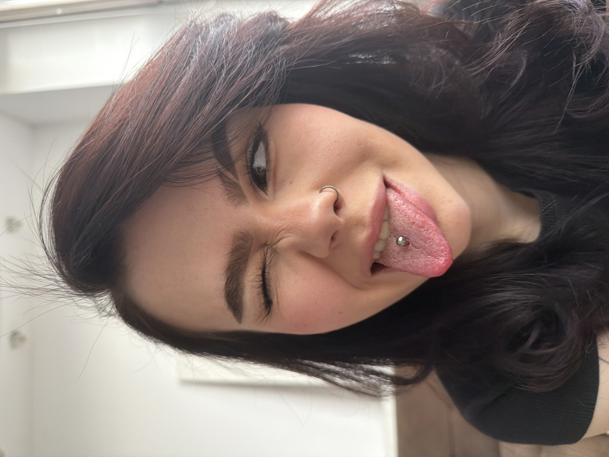 A close-up portrait features a young person, likely female, captured at a dynamic diagonal angle. The subject has dark brown hair that frames their face. They are making a playful expression, with one eye (viewer's right) winking or squinting, and the other eye open, looking directly towards the camera. Their tongue is extended outwards, revealing a silver barbell piercing. A silver hoop nose ring is also prominently visible in their left nostril. The skin appears clear with a hint of natural flush on the cheeks. The subject is wearing a dark-colored top, likely black. The background is softly blurred and light-toned, suggesting an indoor setting with diffused lighting. The overall impression is one of youthfulness, expressiveness, and a hint of mischievousness. No text is visible in the image.